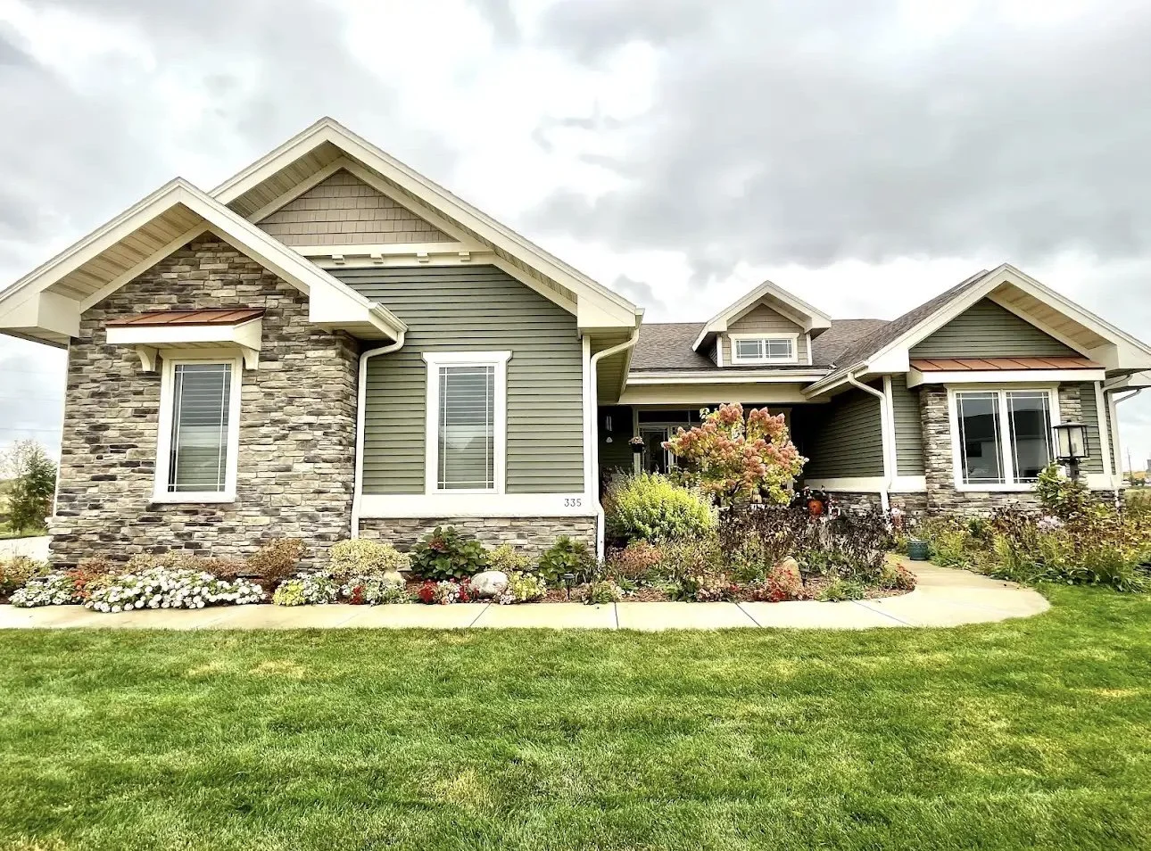 Front view of a modern house with a green exterior, stone accents, and a landscaped front yard under a cloudy sky.