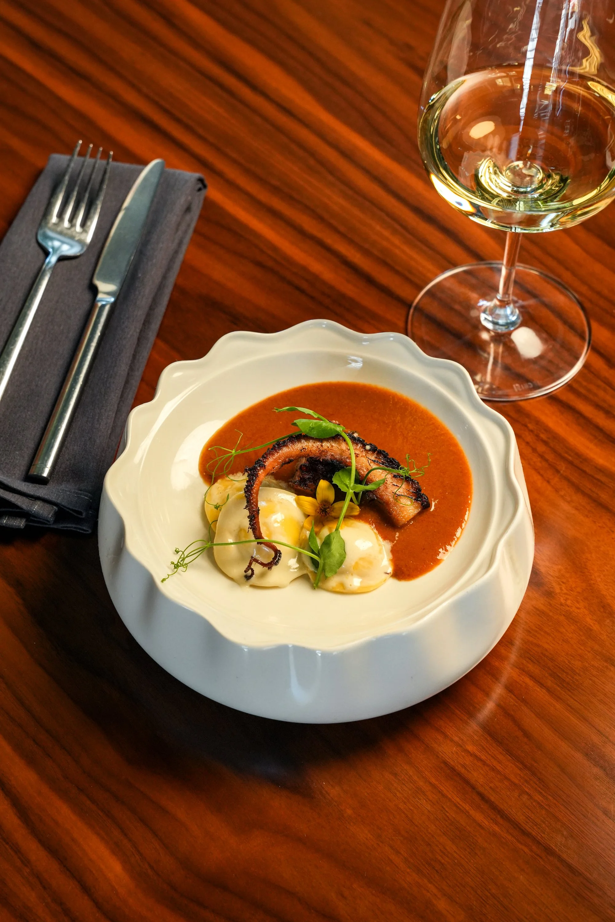 fine dining plate with a piece of octopus in the sauce with three ravioli on the plate alongside a glass of white wine and silverware