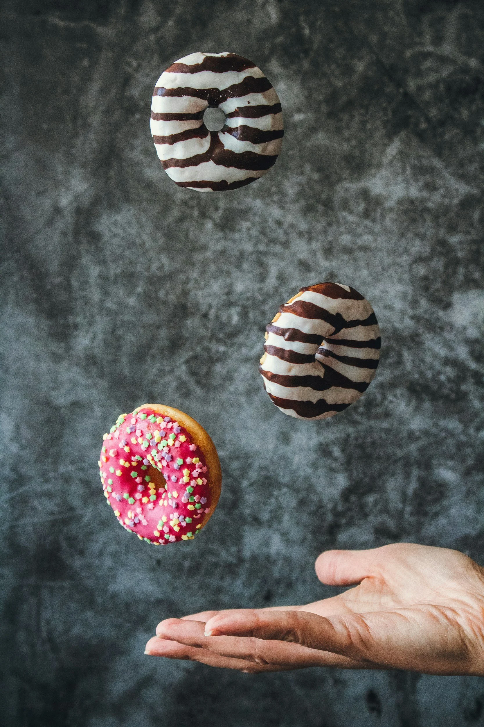 A person's hand is extended with three donuts levitating above it against a gray textured background. Two donuts have white glaze with chocolate stripes, and one has pink glaze with multicolored sprinkles.