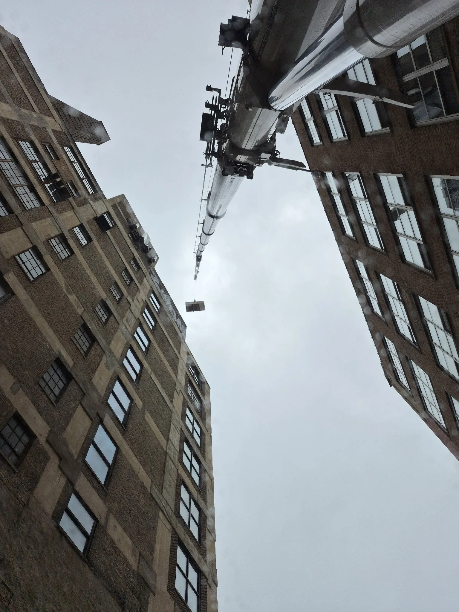 Looking up between two tall brick buildings at a construction crane extending into a cloudy sky.
