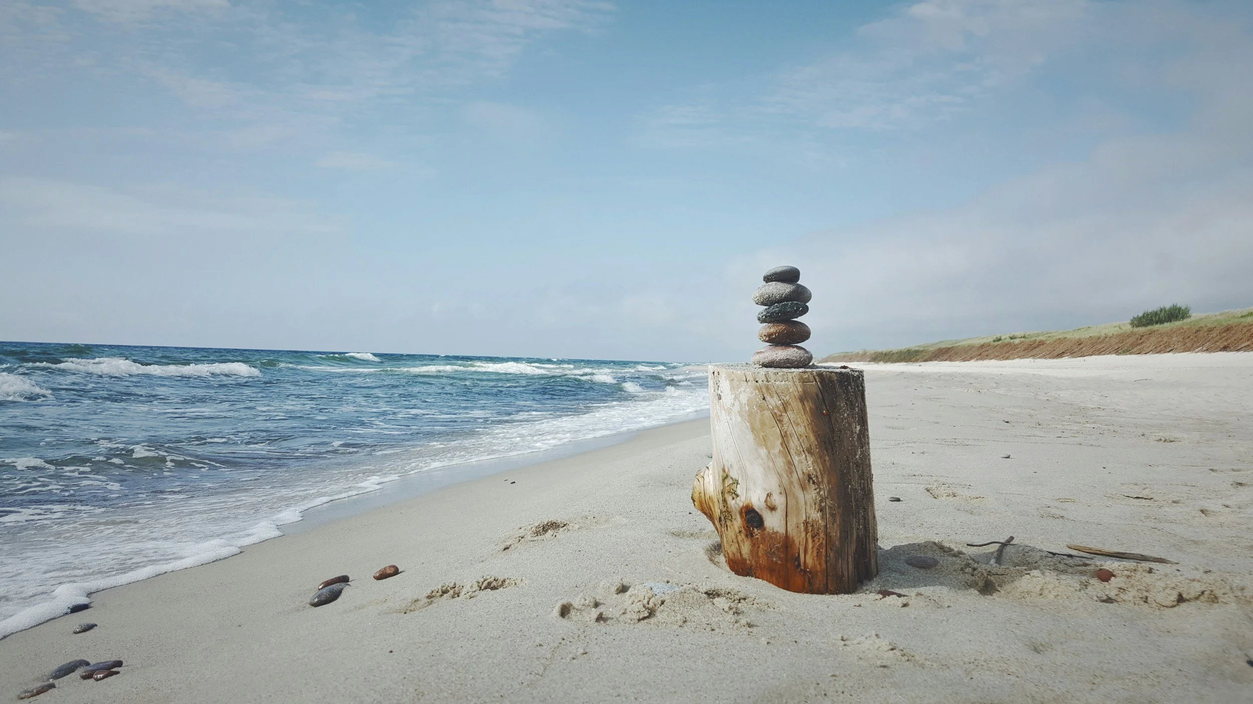 Stacked rocks balanced on a piece of driftwood on a sandy beach with ocean waves and a cloudy sky in the background.