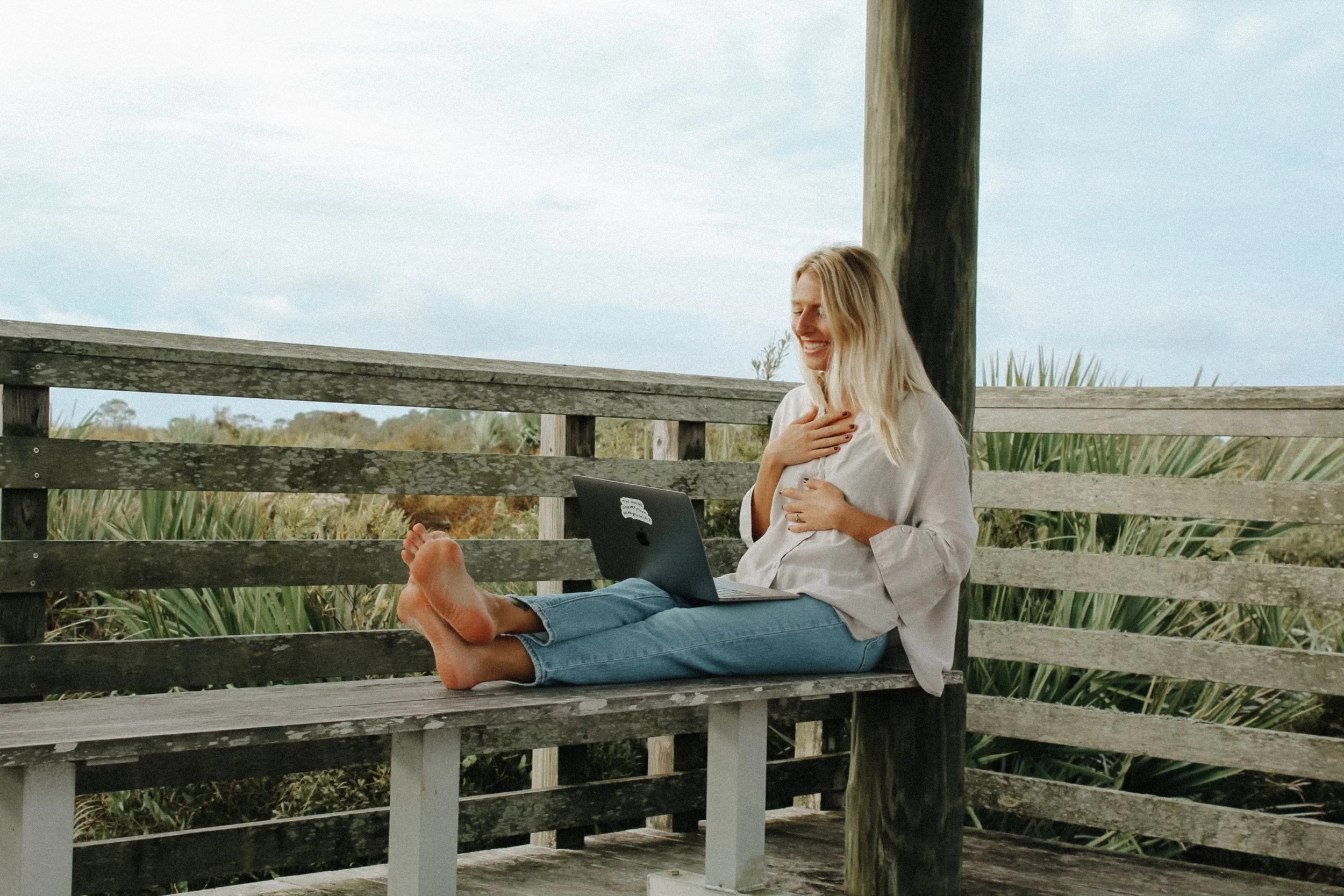 A woman sitting on a wooden bench outdoors, smiling with her hand on her chest, with a laptop on her lap and barefoot, surrounded by greenery and a cloudy sky.