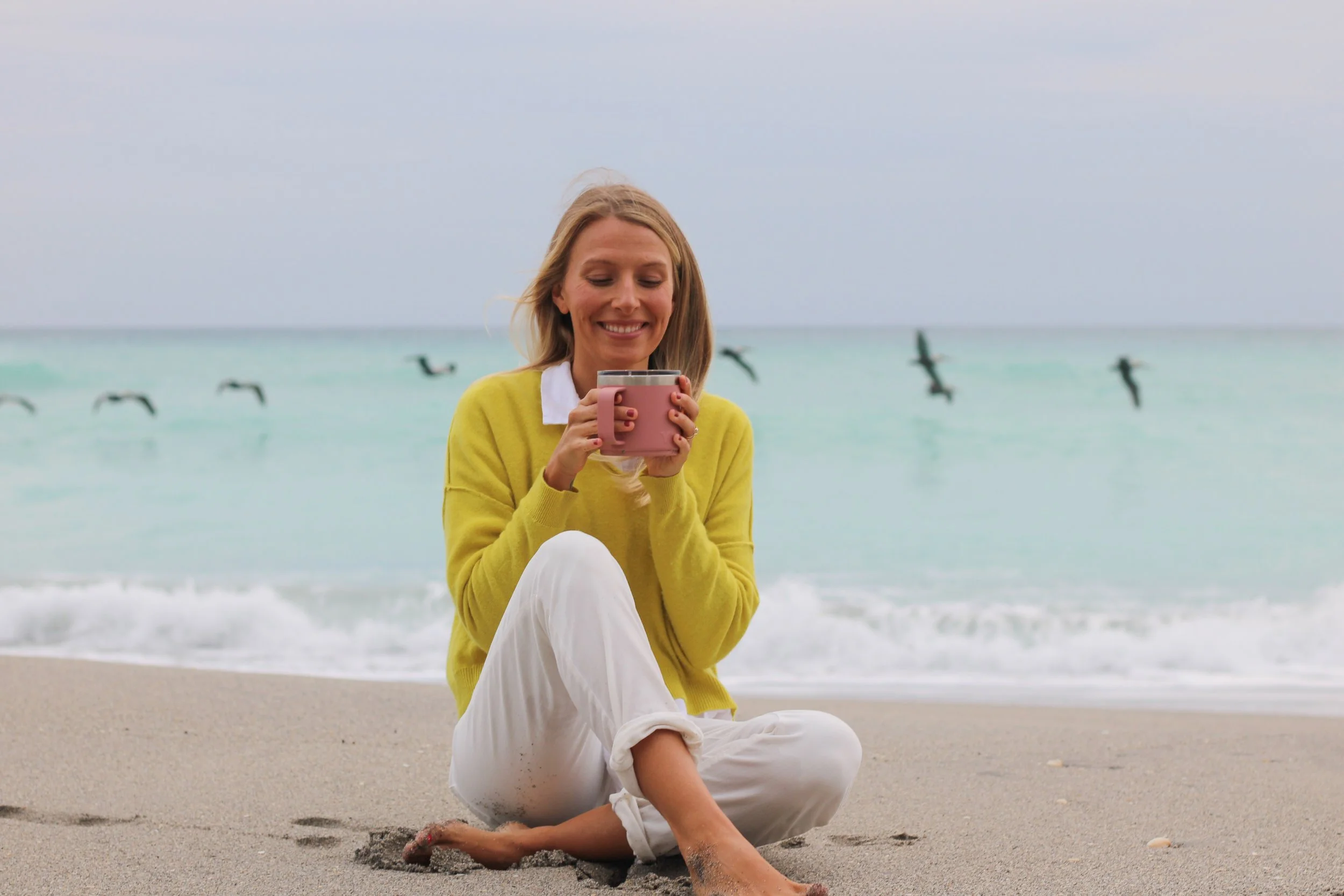 A woman sitting cross-legged on the beach, smiling and holding a pink mug, with seagulls flying over turquoise water in the background.