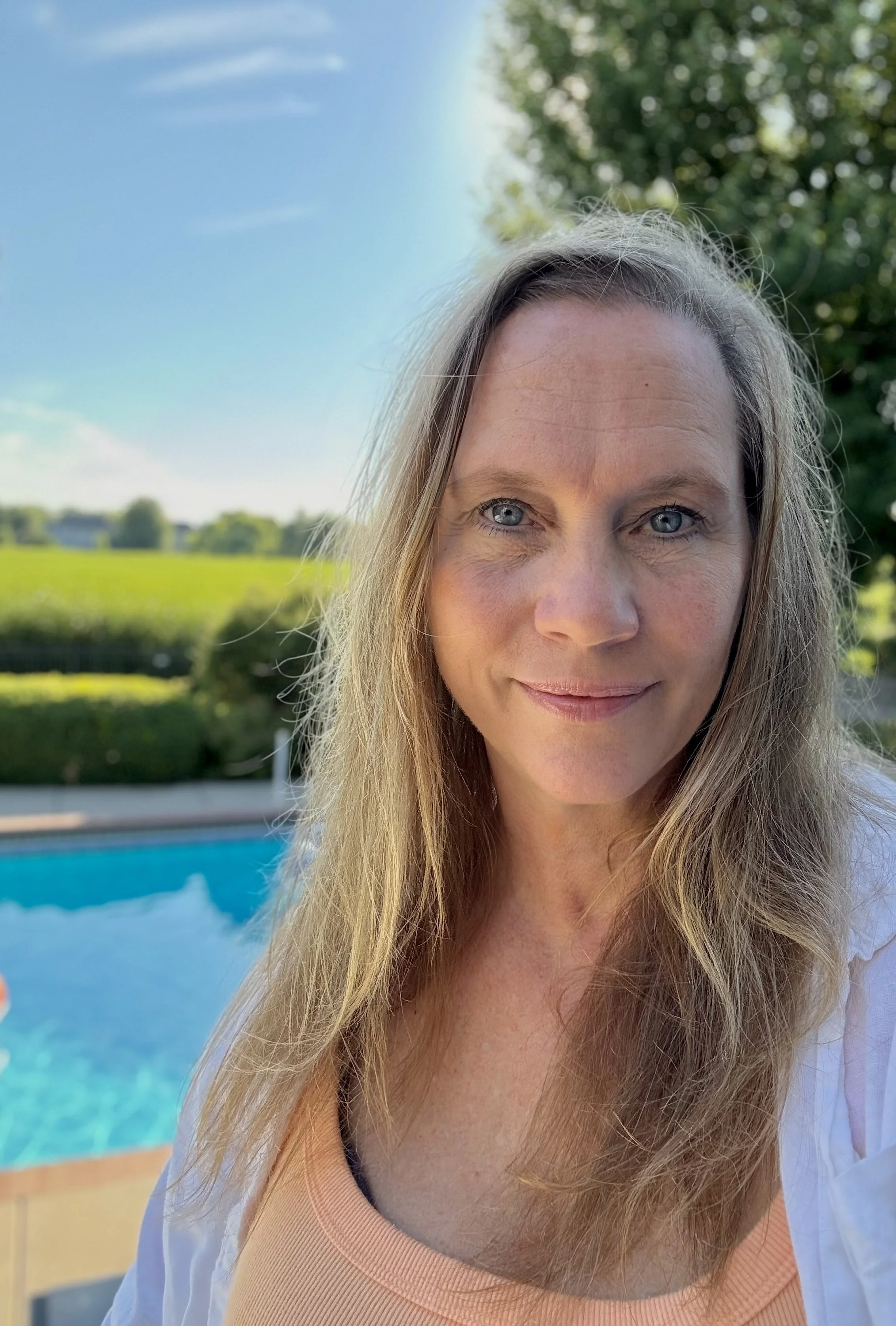 A woman with long blonde hair and blue eyes smiling outdoors by a swimming pool with a green field and trees in the background under a clear blue sky.