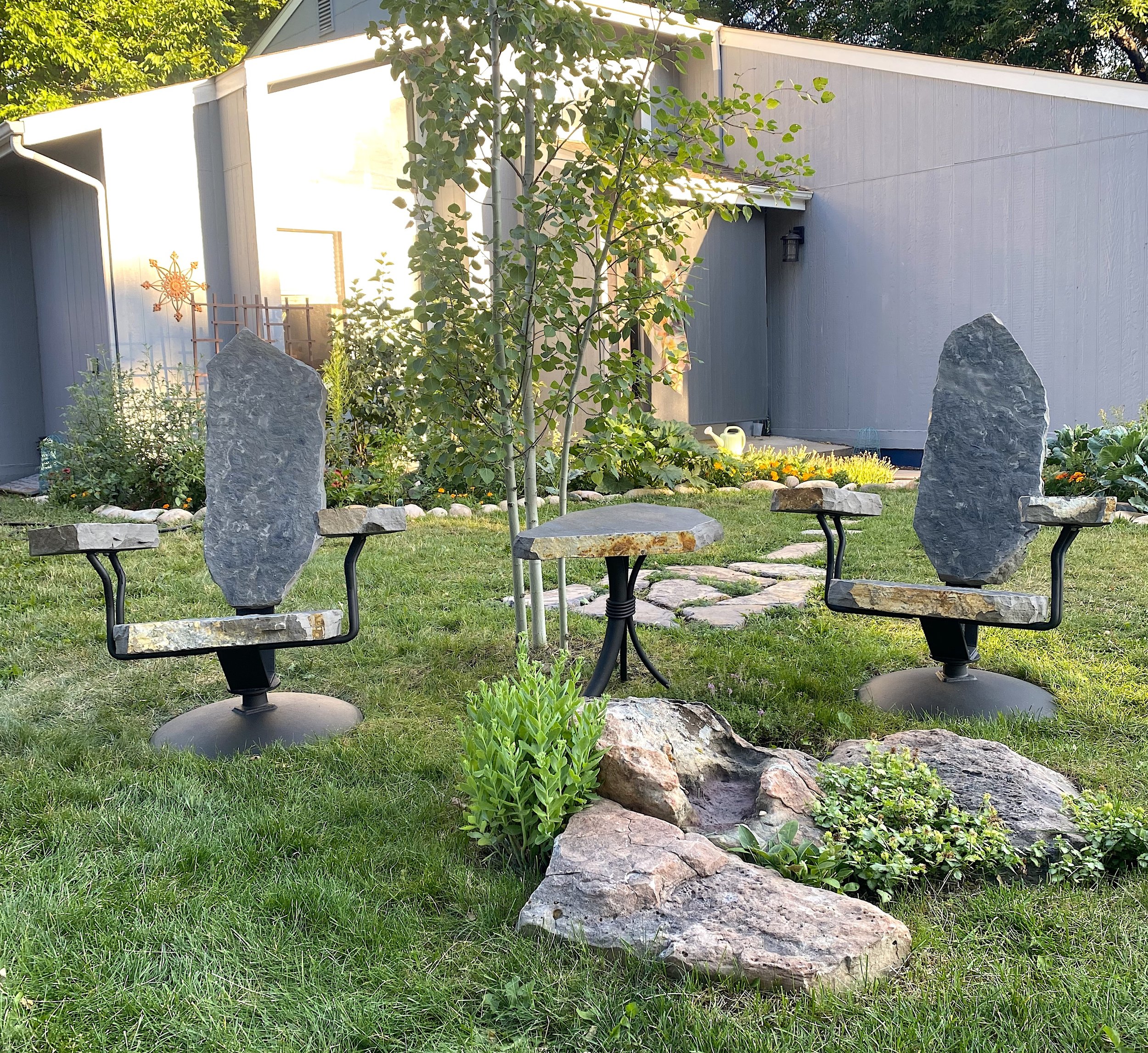 Garden area with two chairs made of large stones and metal frames, a small stone table, green grass, plants, and a gray building in the background.