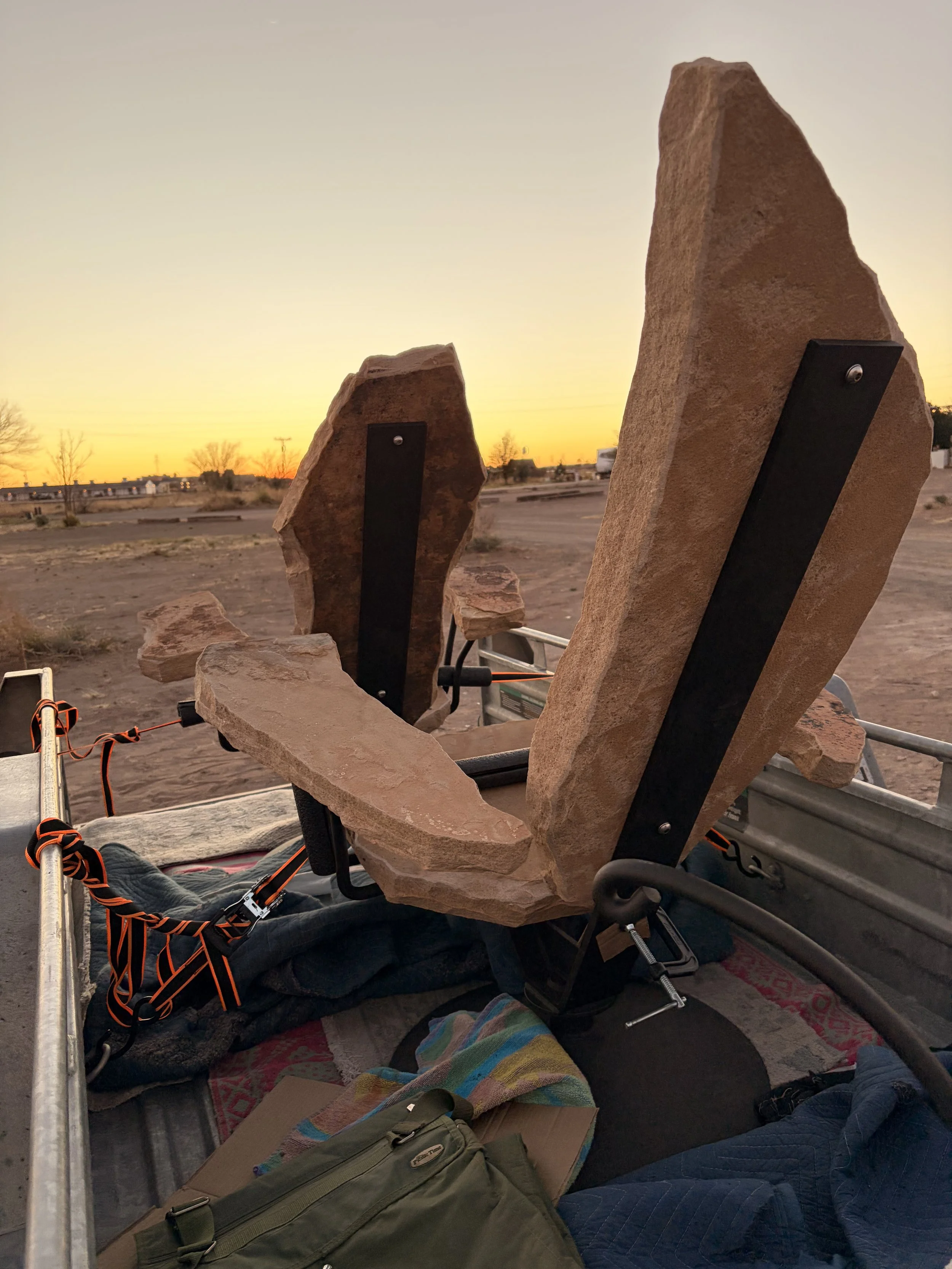 Large reddish-brown stone arranged vertically in a frame, with a smaller stone to the left, set against a sunset sky with a desert landscape and sparse trees in the background.