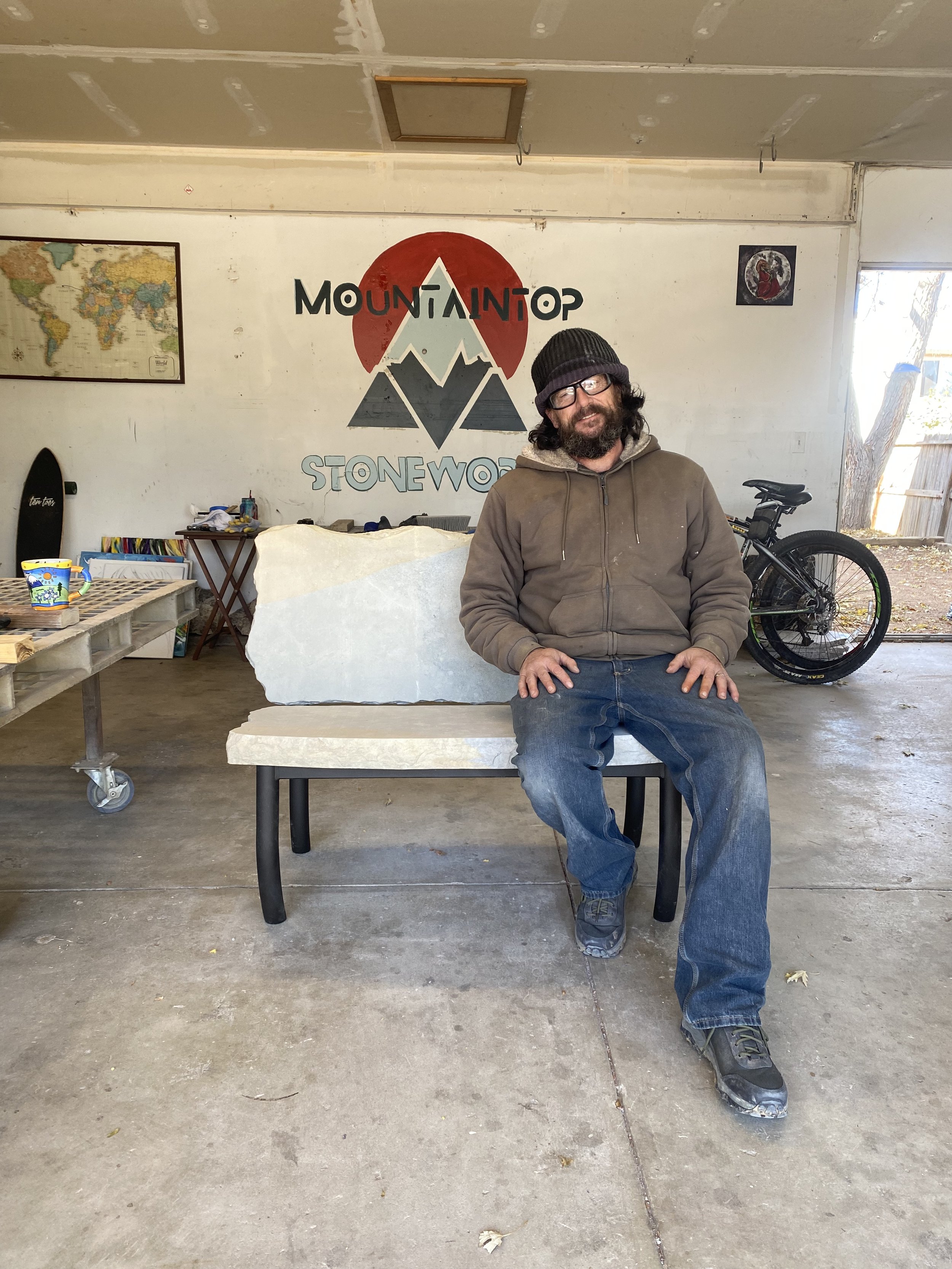 Garry Carmack sitting on stone bench inside a garage with a painted wall that reads 'Mountaintop Stoneworks' featuring a mountain and sun graphic. Bicycles, a world map, and various objects are in the background.