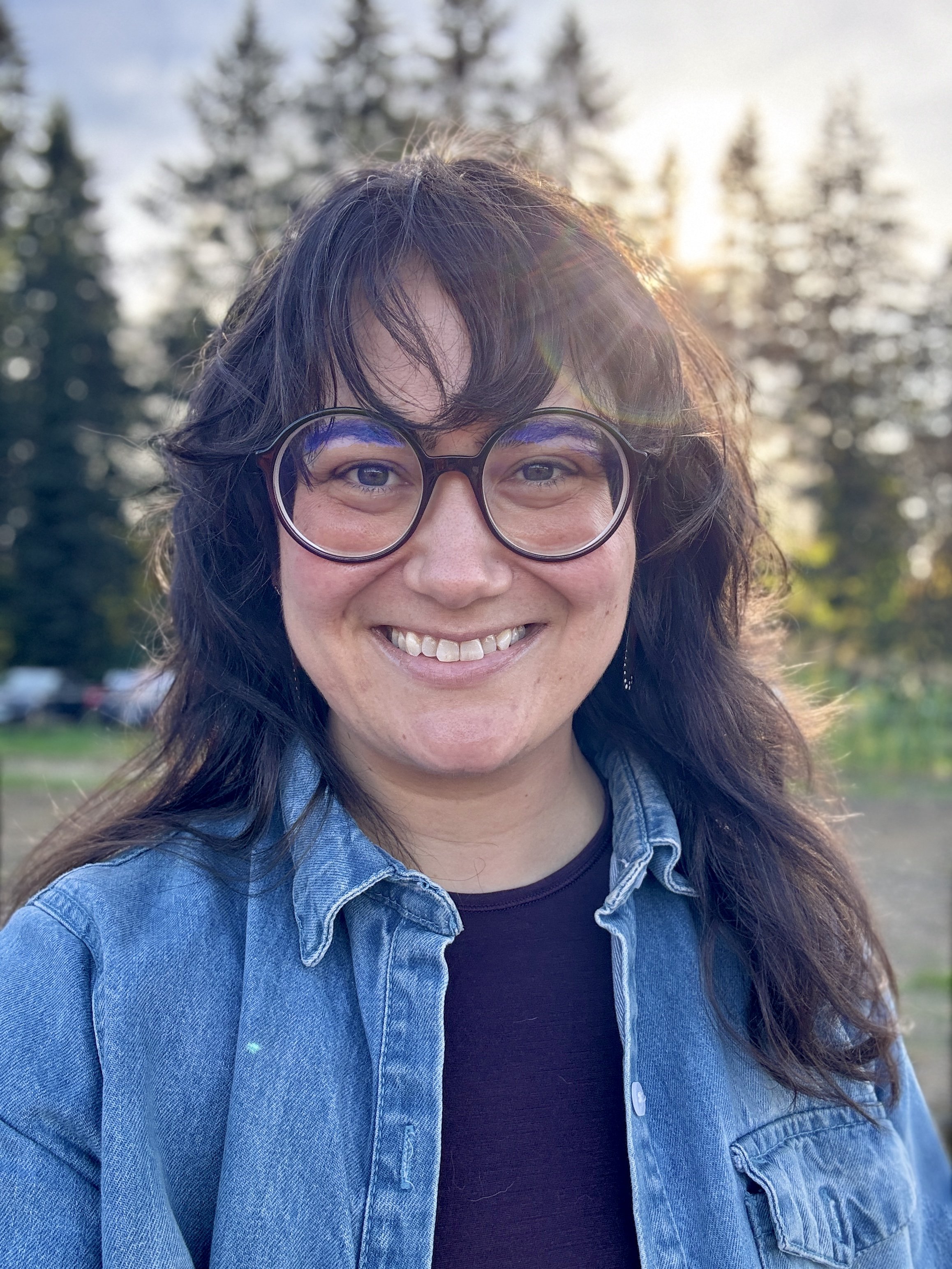 Melody, a white woman with dark shaggy hair and round glasses, stands smiling in front of a row of evergreen trees and the setting sun