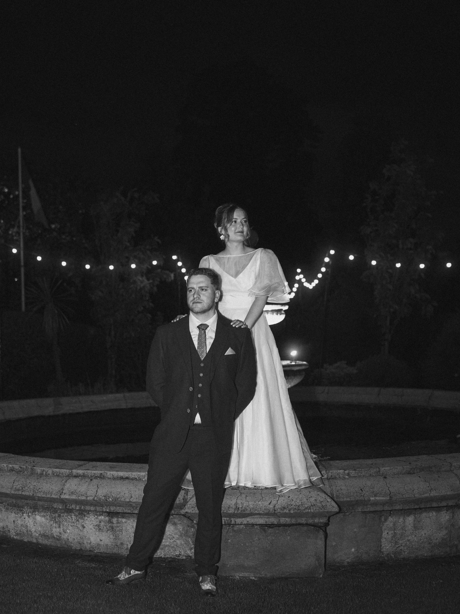 A black and white photo of a man in a suit standing in front of a woman in a wedding dress at night with string lights in the background.