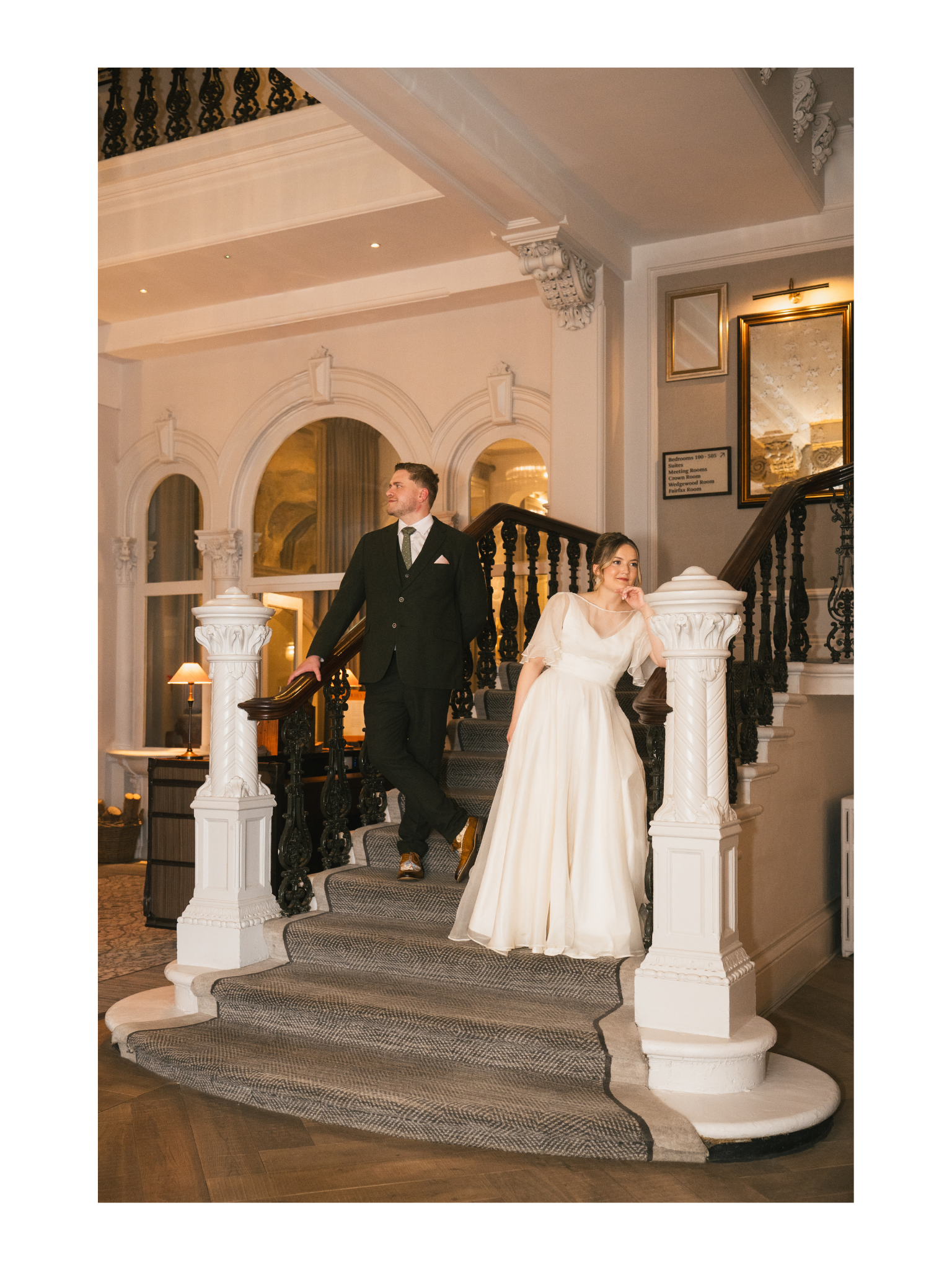 A newlywed couple standing on a grand staircase inside a luxurious hotel or mansion. The groom in a black suit and the bride in a white wedding dress are posing, with the bride leaning on a white ornate pillar and the groom holding the staircase rail