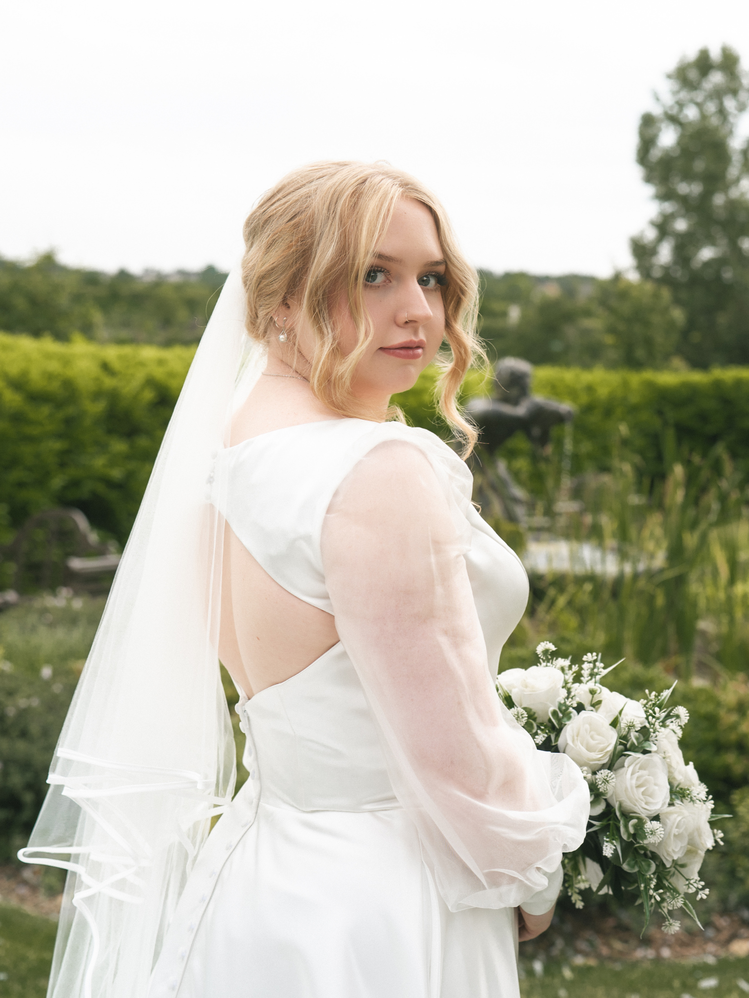 A bride with blonde hair in loose curls, wearing a white wedding dress and veil, holds a bouquet of white roses and greenery outdoors, with trees and greenery in the background.