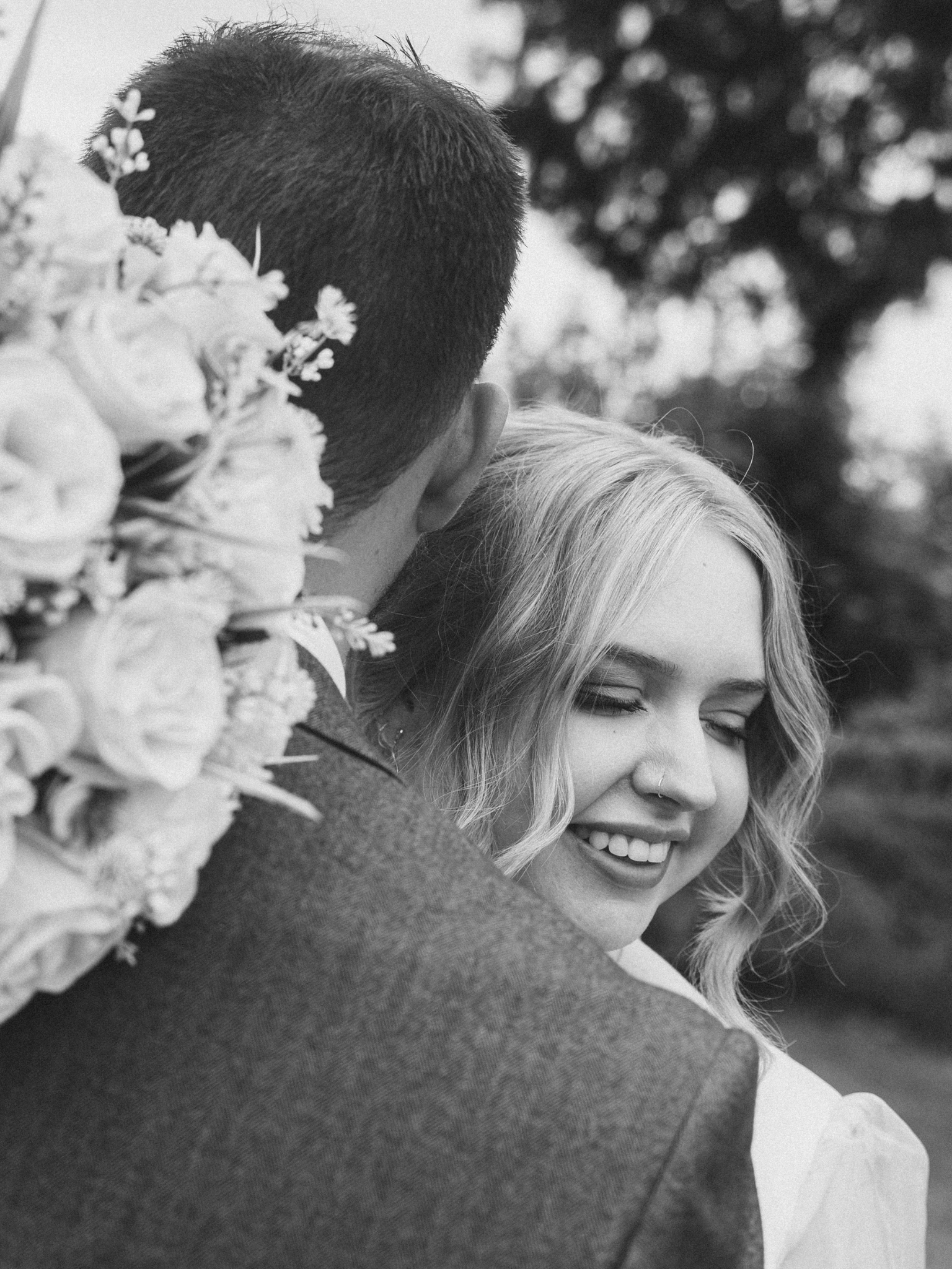 A man with short dark hair and a woman with blonde hair are close together, smiling, with the woman's eyes closed. There is a bouquet of flowers nearby, and the background is blurred with trees.