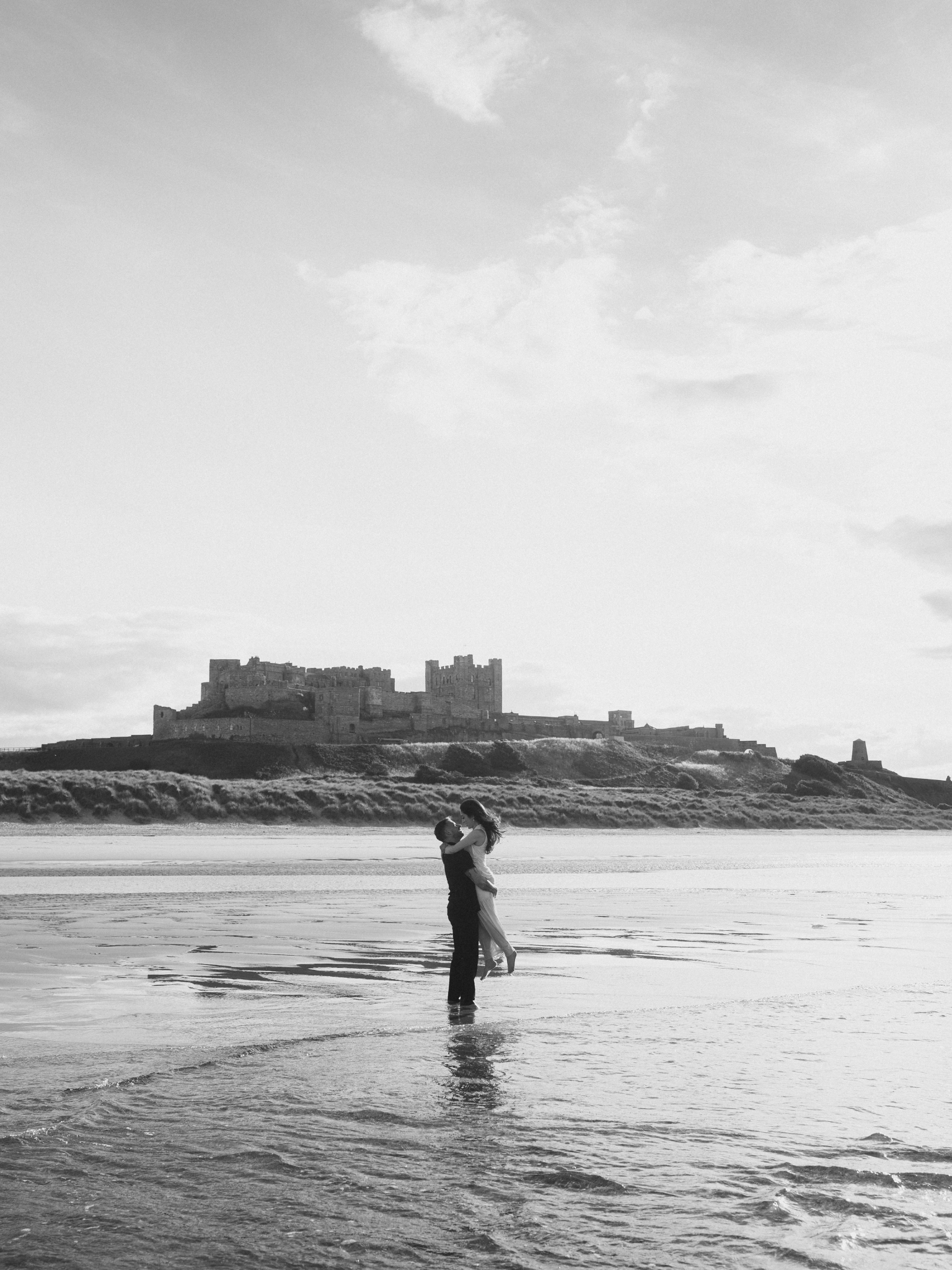 A couple standing in shallow water on a beach, embracing and looking at each other, with a castle on a hill in the background and clouds in the sky.