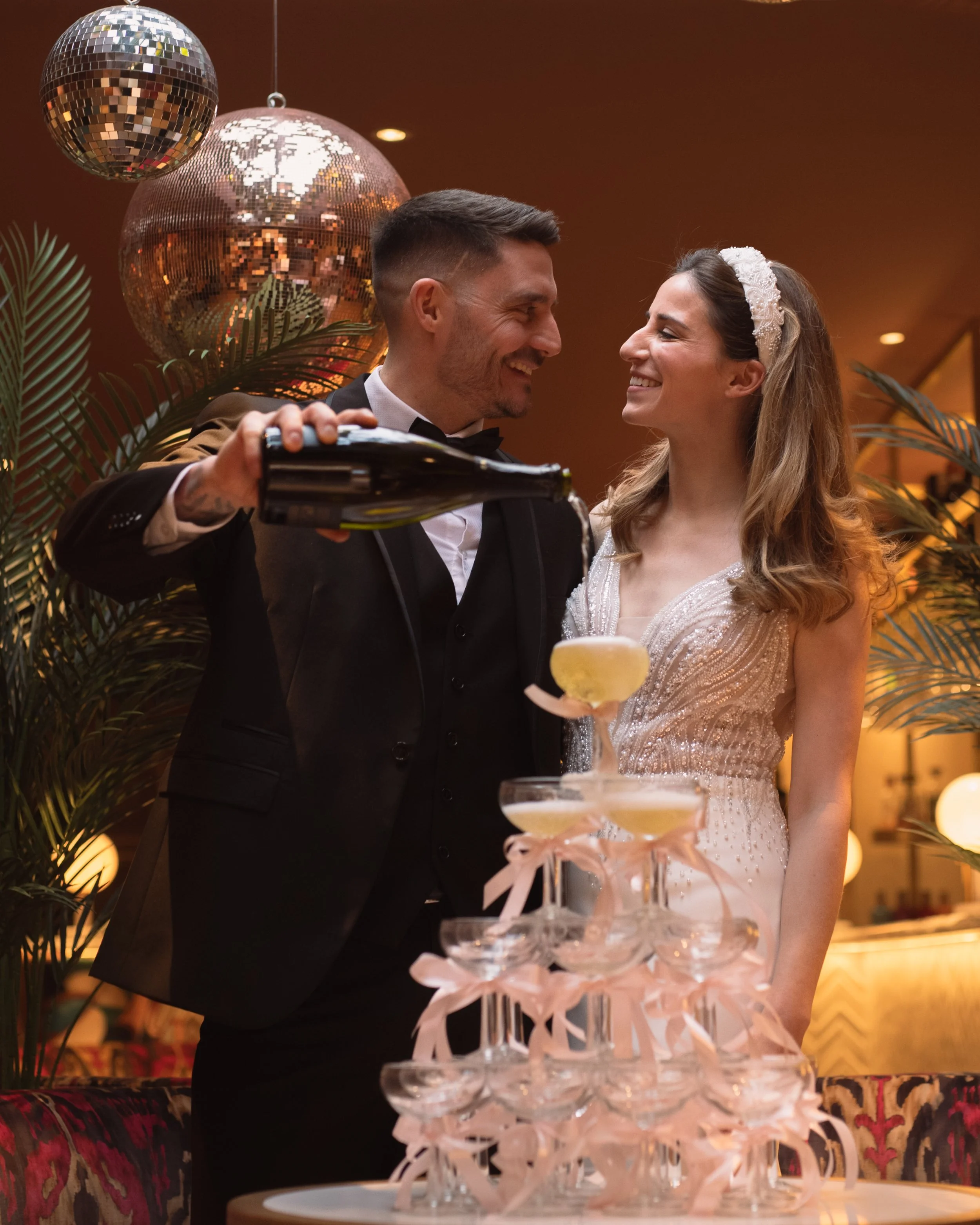 Bride and groom pouring champagne into a tower of glasses beneath disco balls.