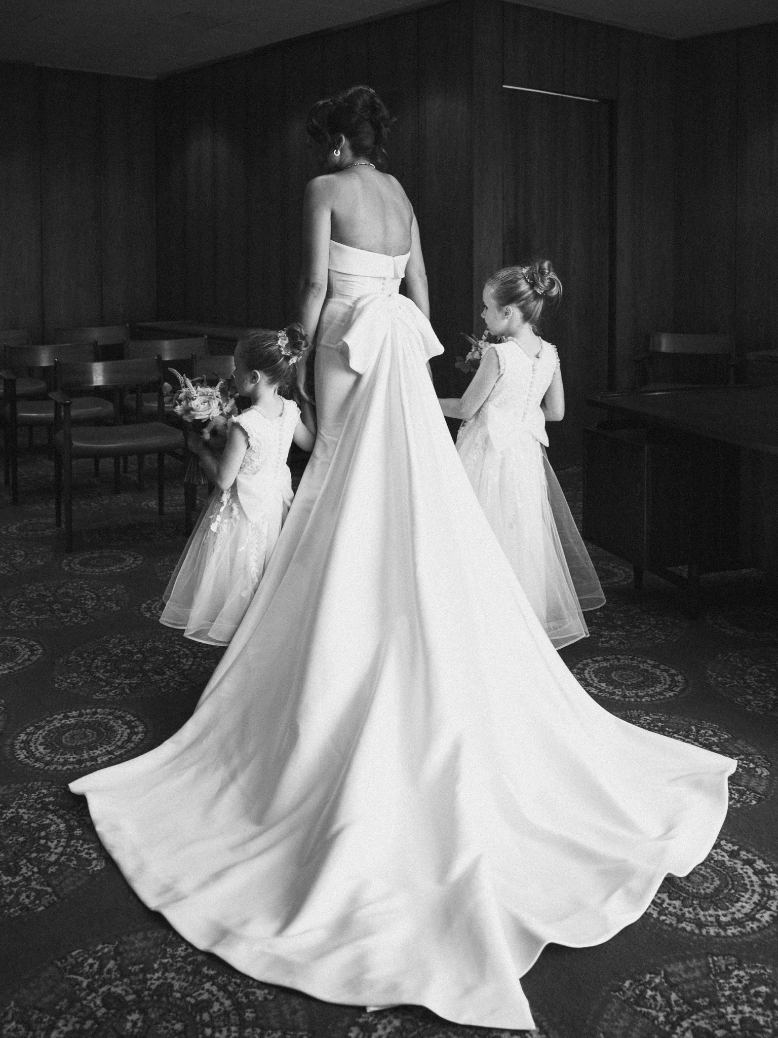 A black and white image of a bride in a strapless wedding dress with a long train standing in a room with wood-paneled walls. She is holding the hand of a young girl in a dress, and another young girl in a similar dress stands nearby, looking at the 