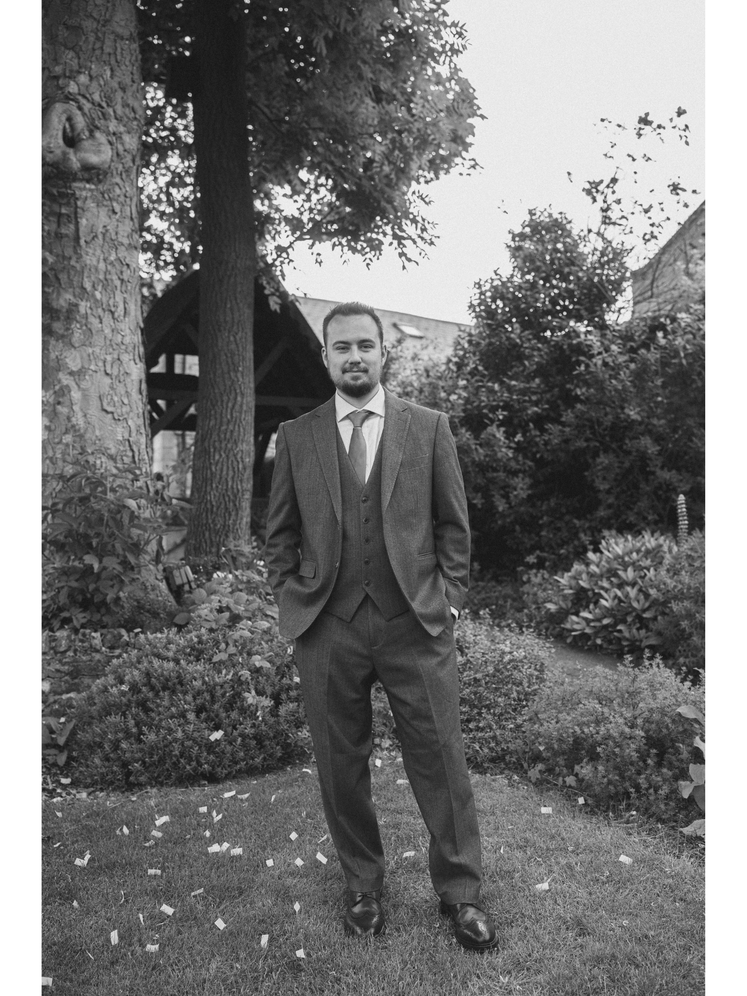A man in a suit standing outdoors in a garden with trees and bushes, with small pieces of confetti scattered on the ground.