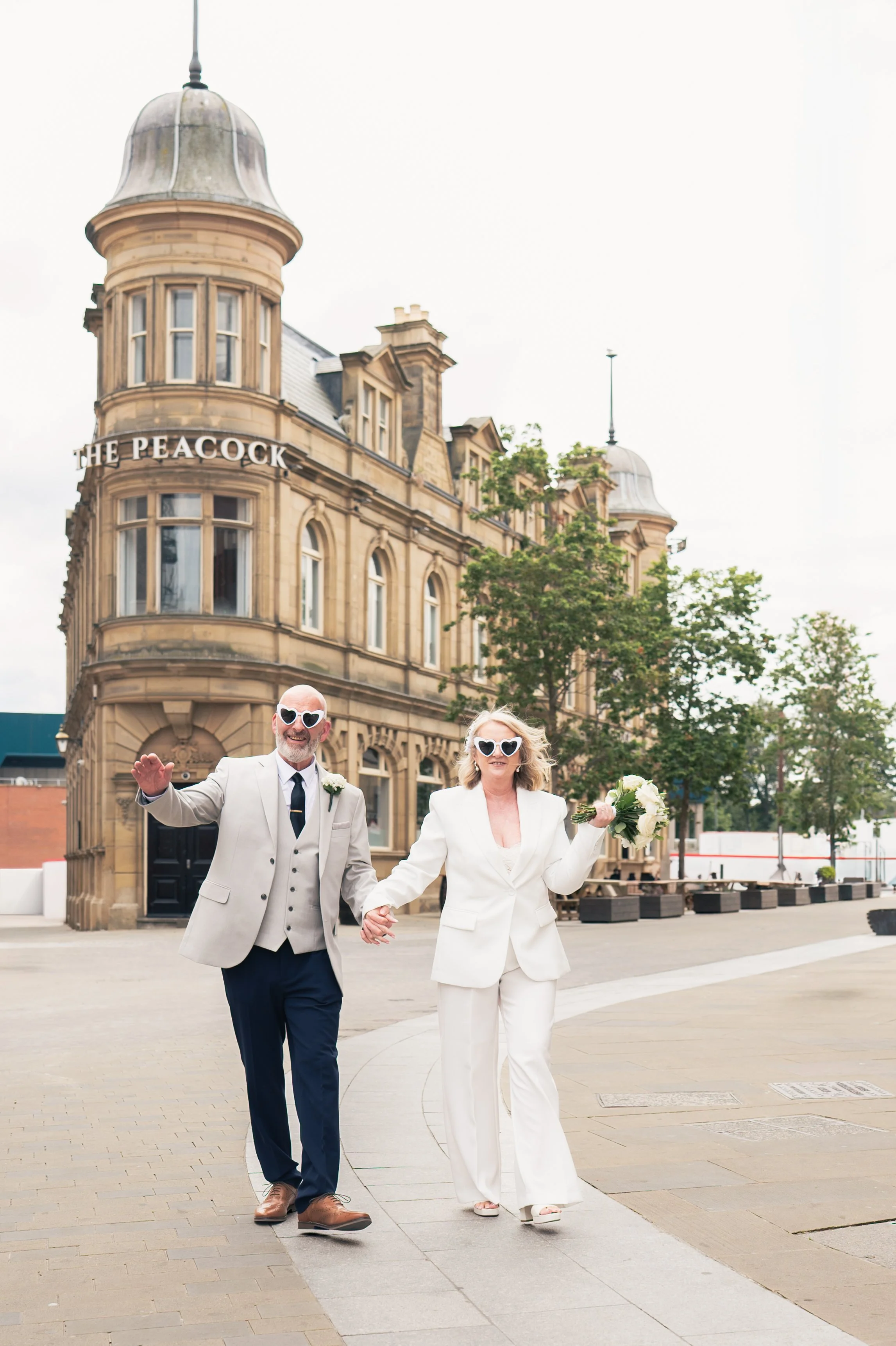 A couple dressed in wedding attire walking hand in hand outdoors near a historic building with the sign 'The Peacock'. They are wearing white sunglasses and smiling, with the woman holding a bouquet of flowers.