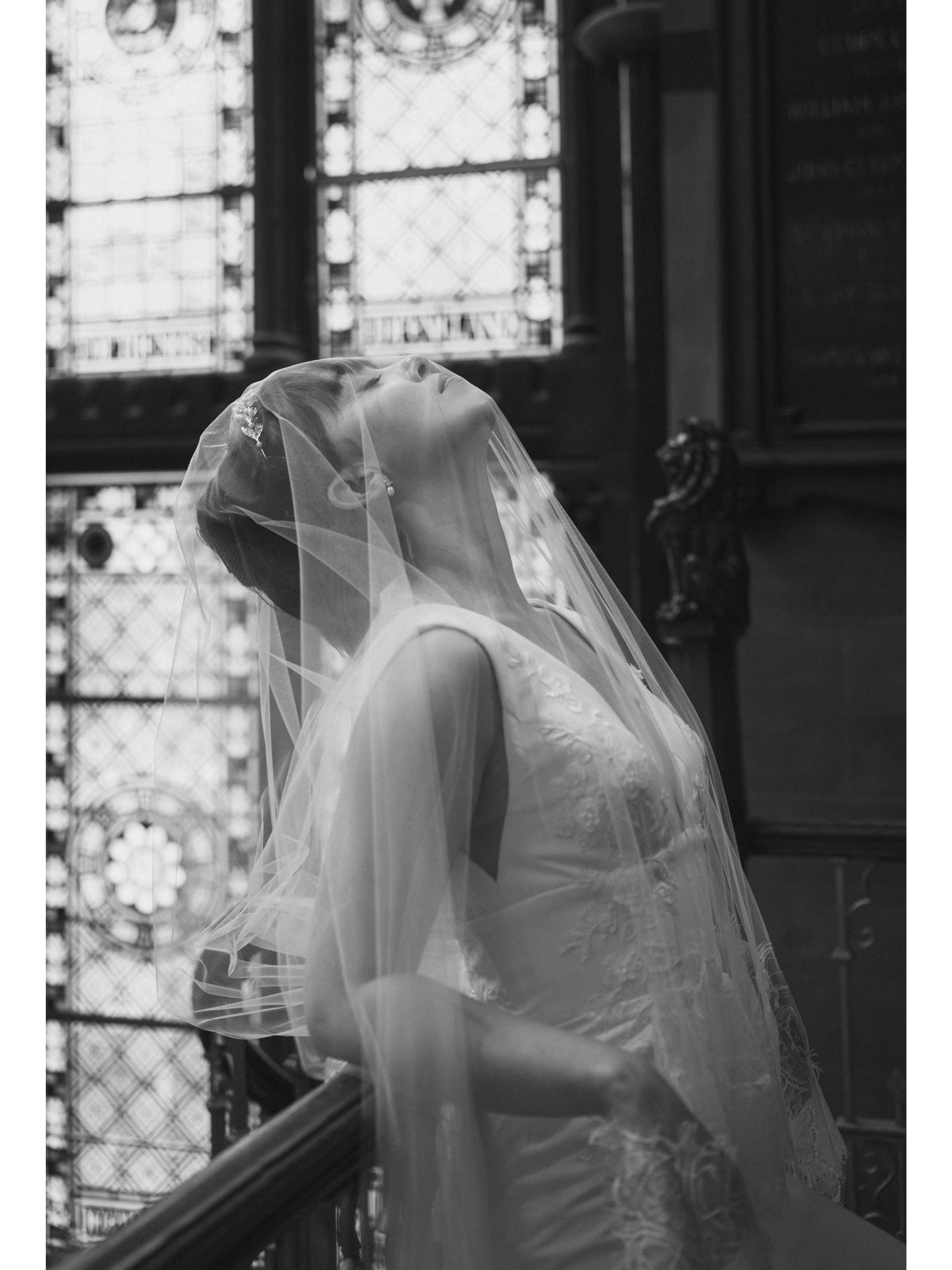 A bride in a wedding dress with a veil looking upward inside a building with ornate stained glass windows.