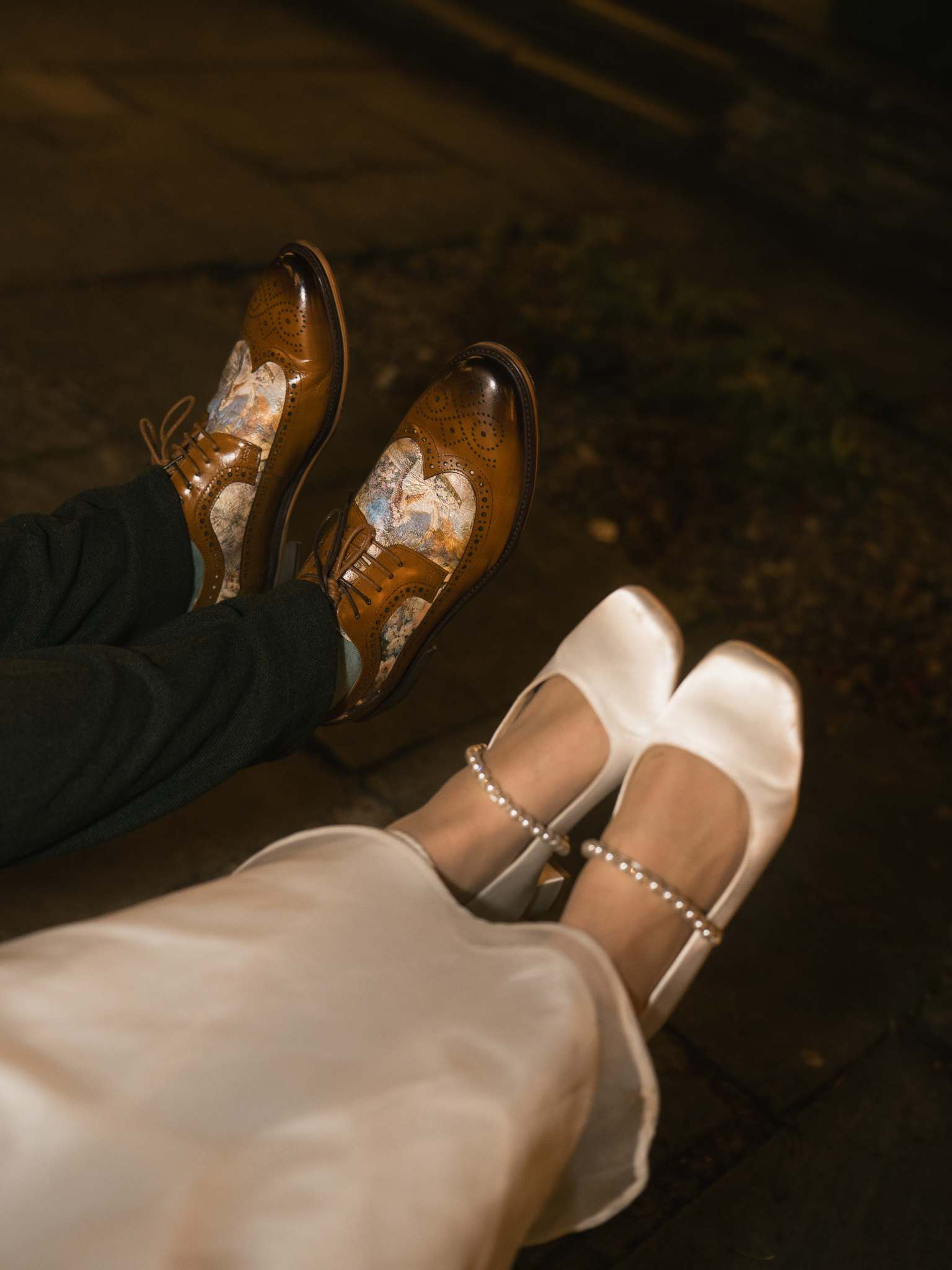 View of two pairs of shoes: one pair of men's polished brown leather dress shoes with intricate pattern, and one pair of women's white satin heels with ankle pearl anklets, worn by two seated individuals.