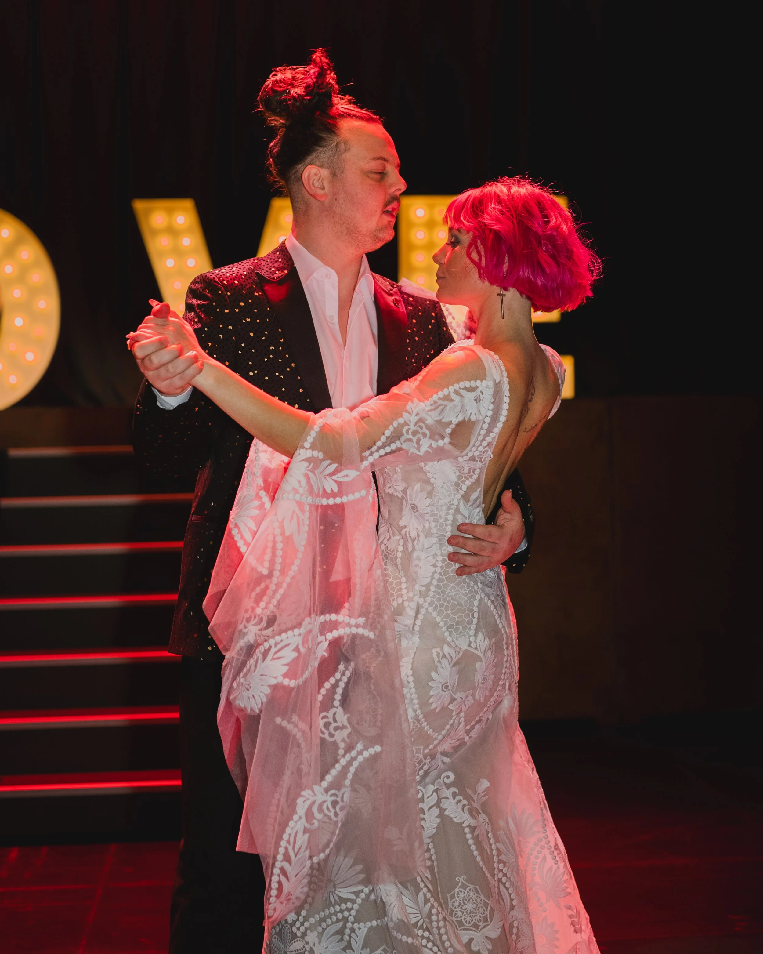Couple dancing in formal attire under stage lights, man wearing a sparkly jacket, woman in a lace dress.