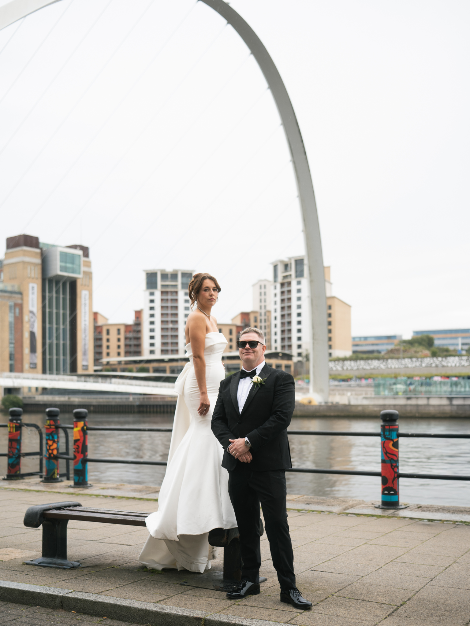 A bride and groom pose on a riverside promenade with modern city buildings in the background on an overcast day.