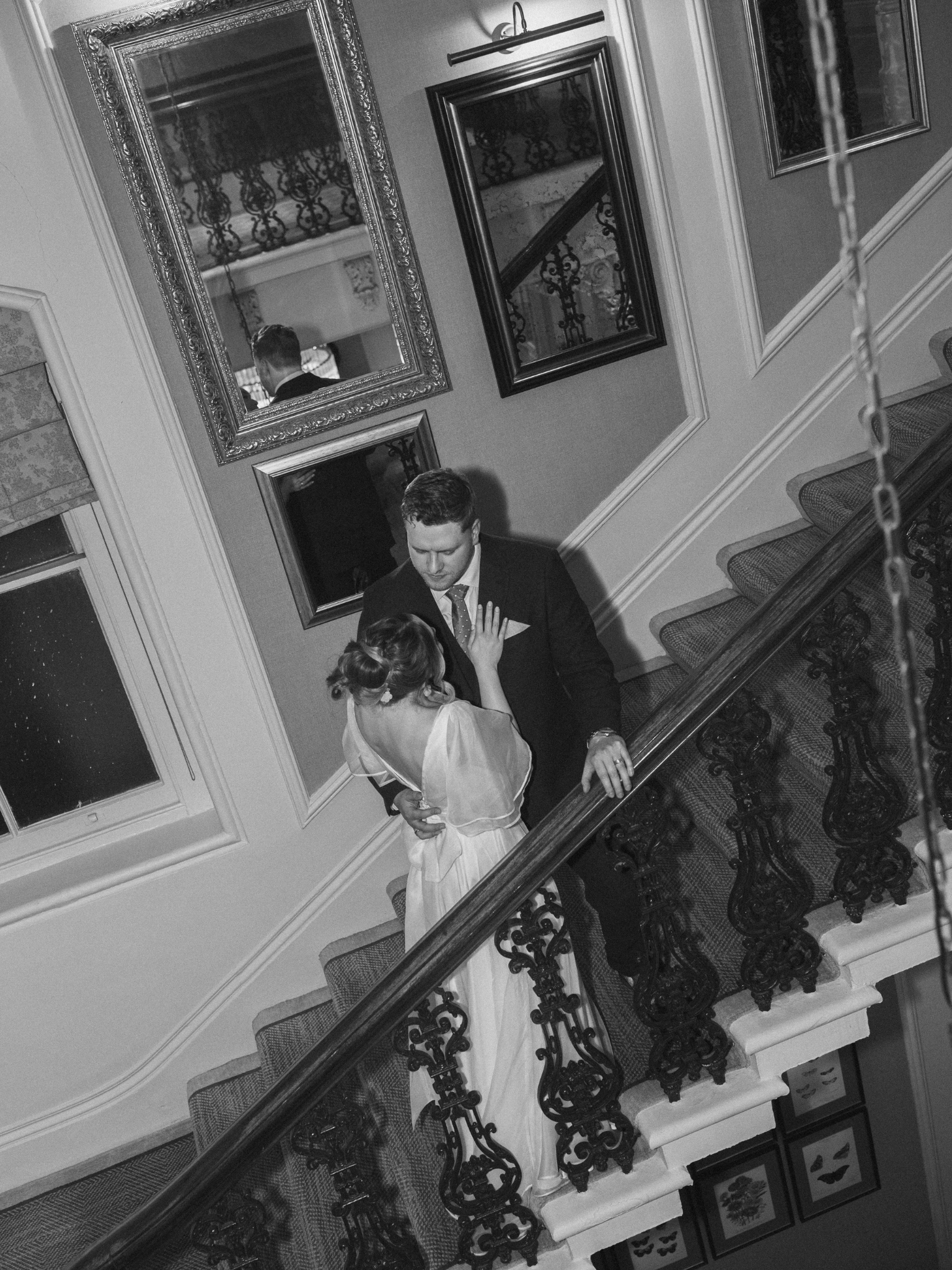 A black and white photo of a man and woman dancing on a staircase, with mirror reflections showing the woman from the back. The staircase has ornate iron railings, and there are framed pictures on the wall alongside multiple mirrors reflecting parts 