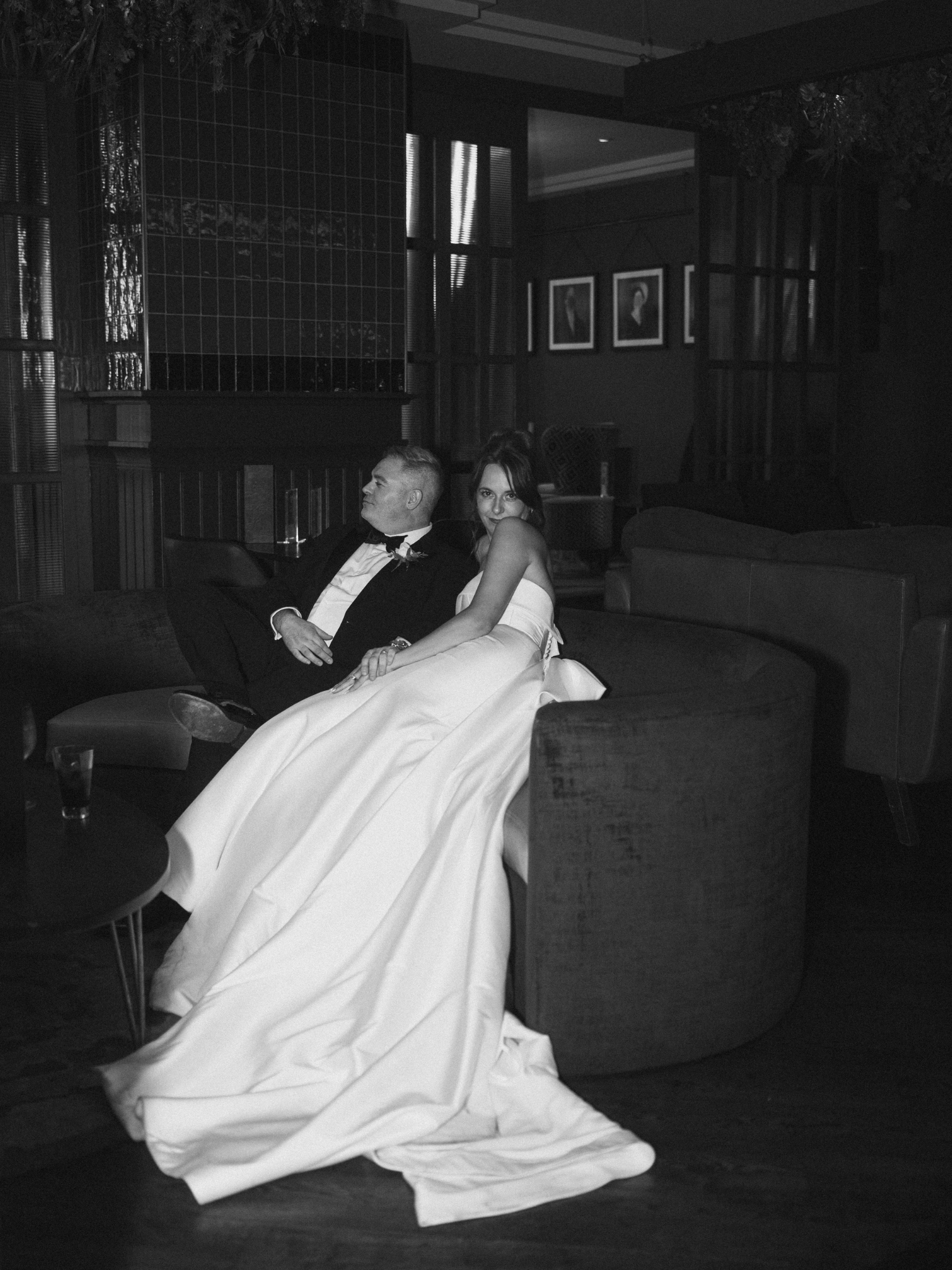 A black and white photo of a woman in a white dress and a man in a tuxedo, sitting on a couch in a dimly lit room, with framed pictures on the wall behind them.