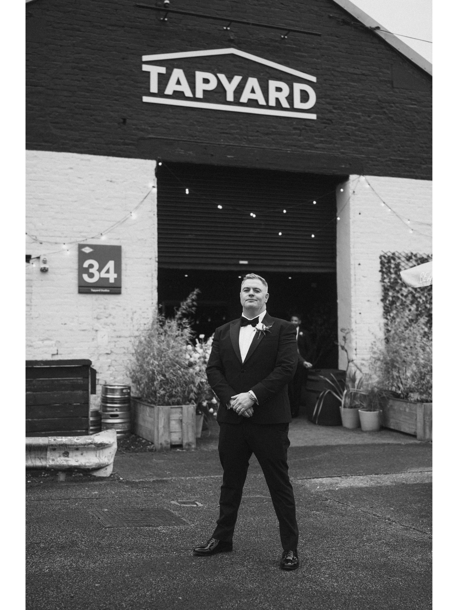 A man in a tuxedo and dress shoes standing outside in front of a building with the sign 'TAPIYARD' on it, surrounded by potted plants and decorative planters.