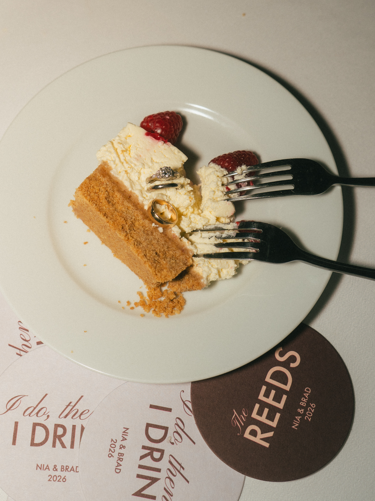 A white plate with a slice of cheesecake, raspberries, and a silver ring with small diamonds on top of the cheesecake. Two wedding bands are also on the plate, one gold and one silver. Two round flyers or cards with text are underneath the plate, par