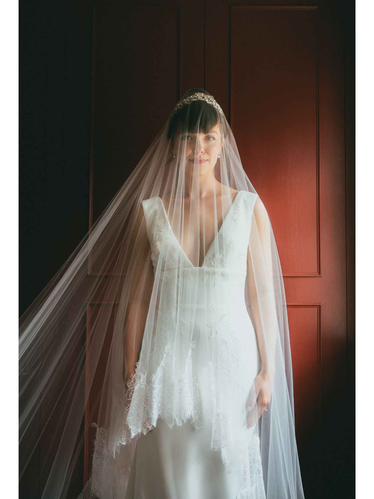 A woman in a wedding dress and veil stands in front of a wooden panel wall, smiling softly.