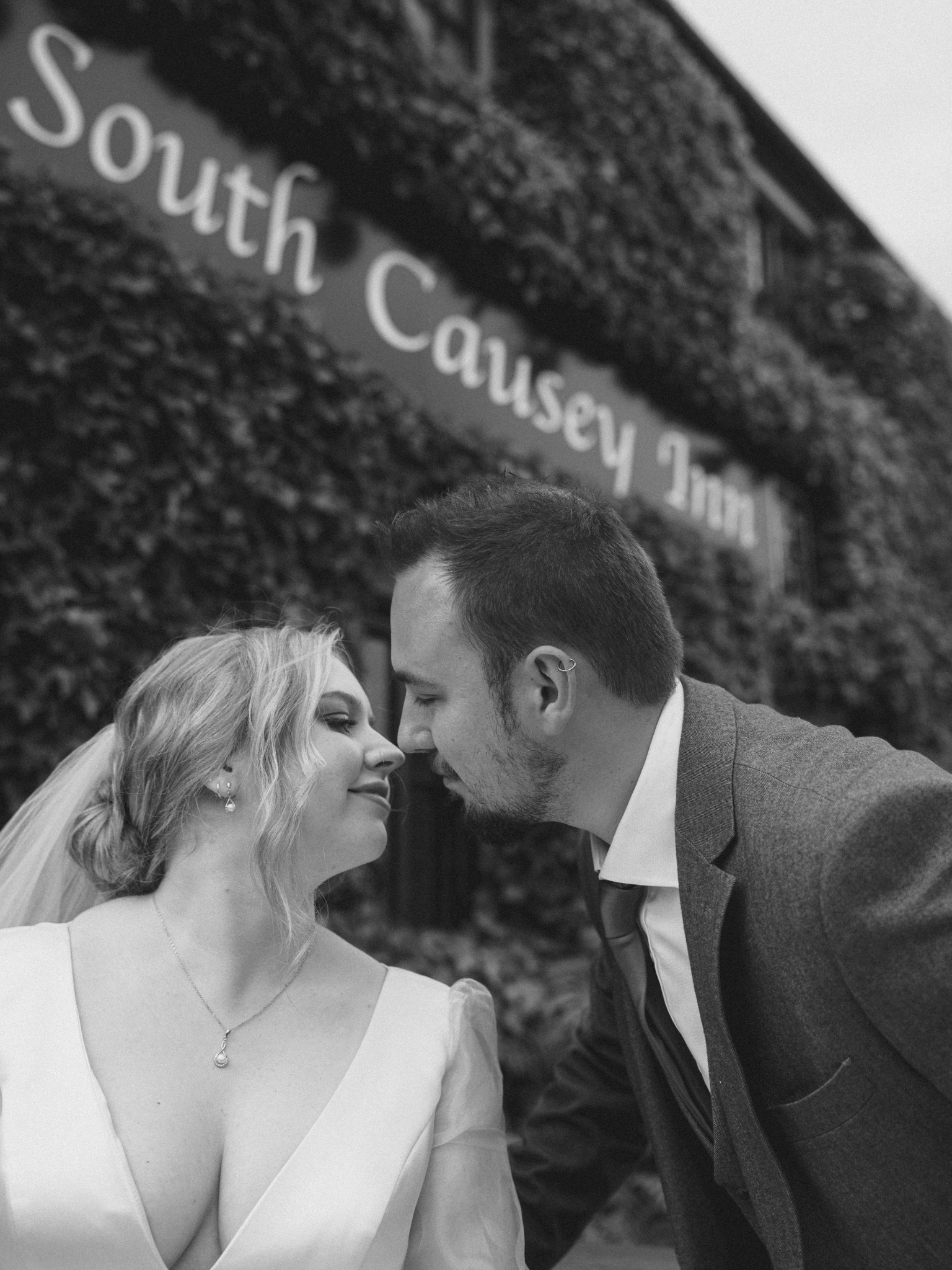 A black and white photo of a bride and groom facing each other closely, about to kiss. The bride has blonde hair, wears a white dress with a deep neckline, and a pendant necklace. The groom has short hair, facial hair, and wears a suit. There is a si