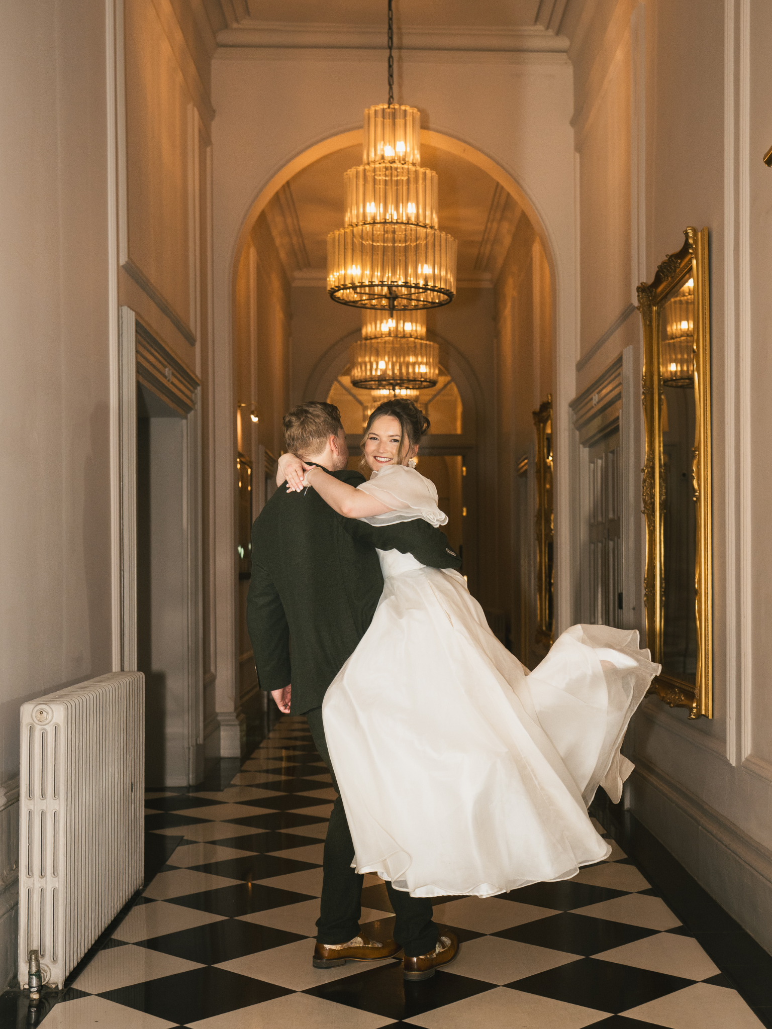 A man in a tuxedo holding a woman in a wedding gown as they dance or embrace in a hallway with black and white tiled floor, chandeliers, and large mirrors.