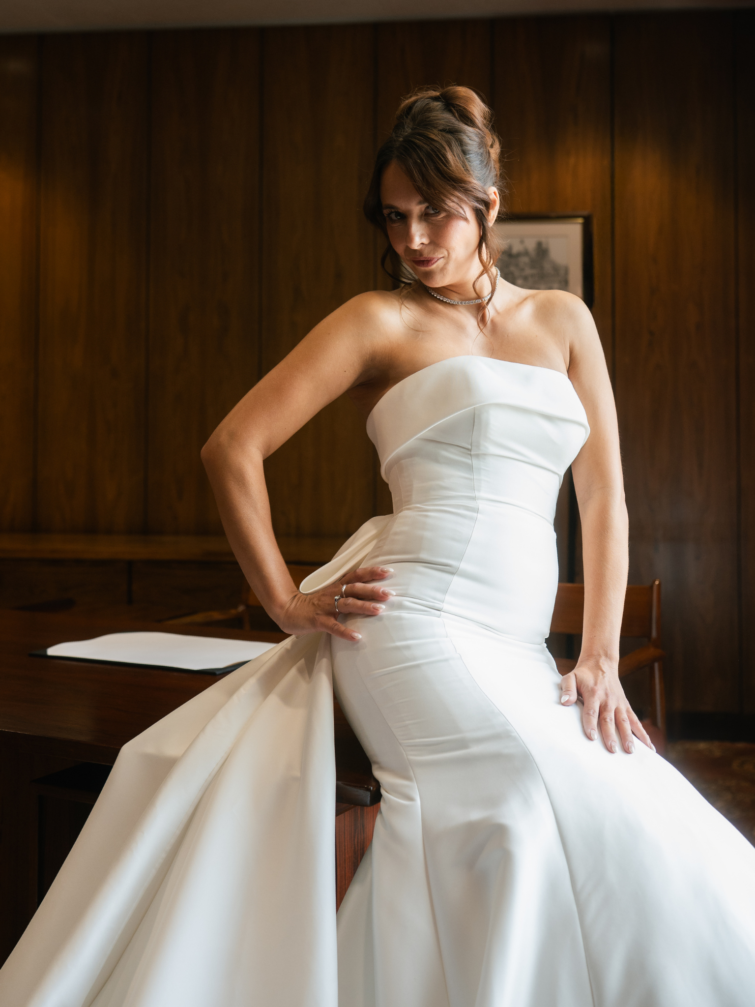 A woman wearing a white strapless wedding dress, posing confidently with one hand on her hip in an indoor setting with wood paneling and a picture frame in the background.