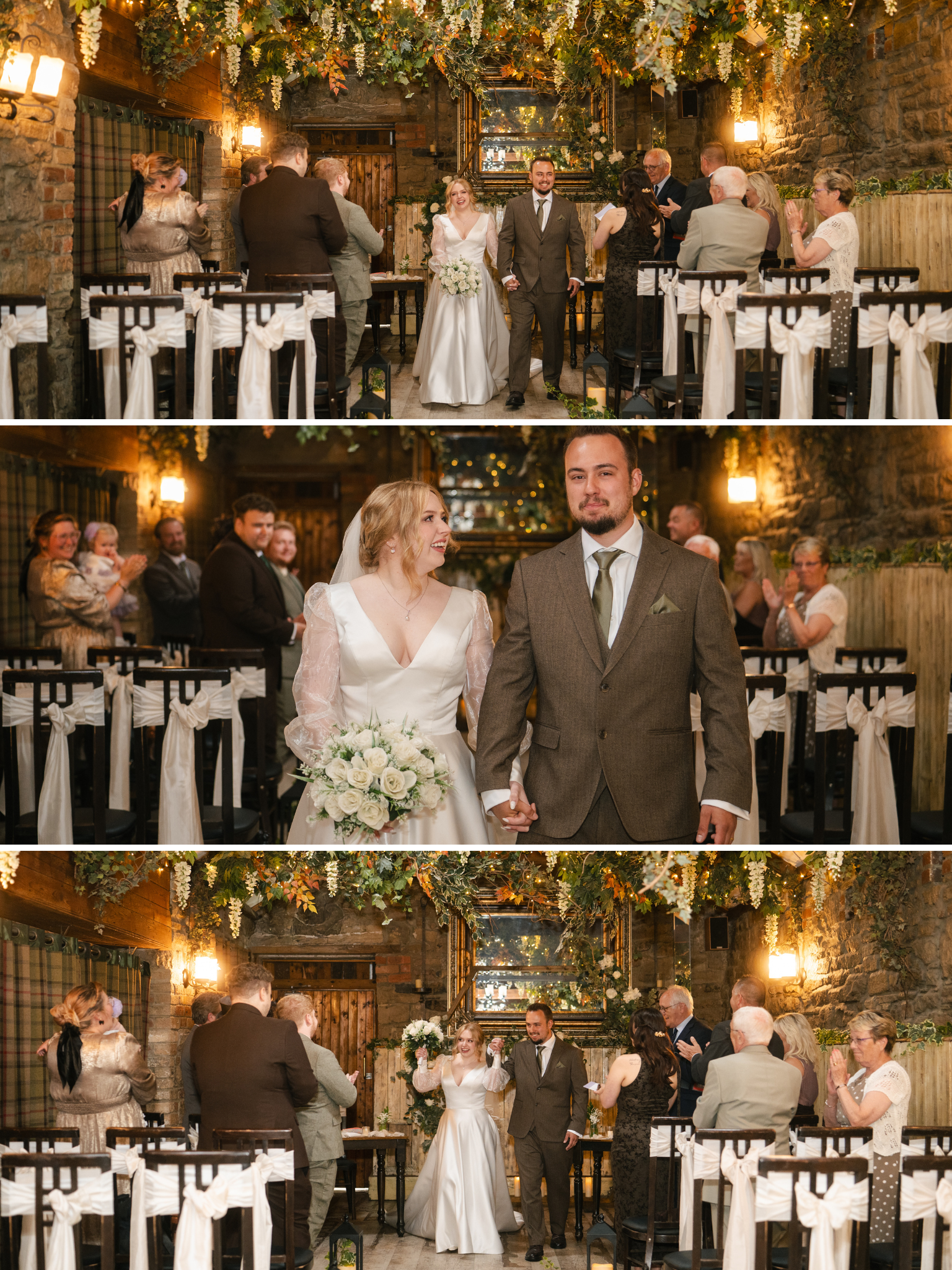 A wedding ceremony with the bride and groom holding hands and smiling, surrounded by guests seated at decorated tables in a rustic, warmly lit venue with floral and greenery decorations overhead.