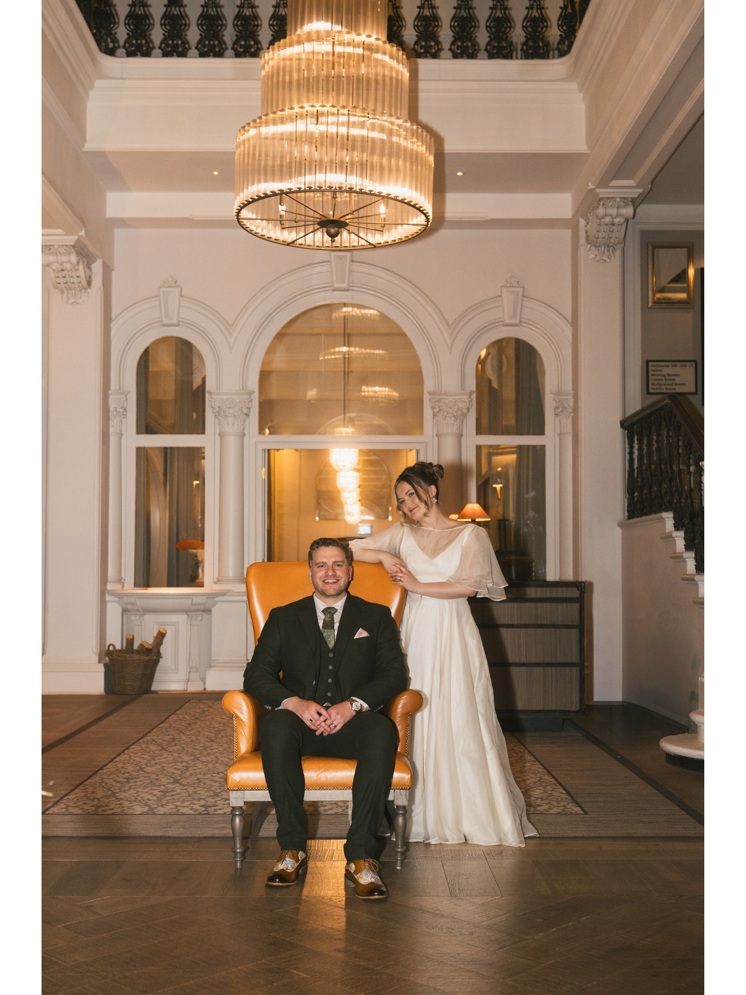 A man in a black suit seated in a yellow armchair with a woman in a white dress standing beside him, holding his hand, in an elegant room with large mirrors, ornate architecture, and a large chandelier hanging from the ceiling.