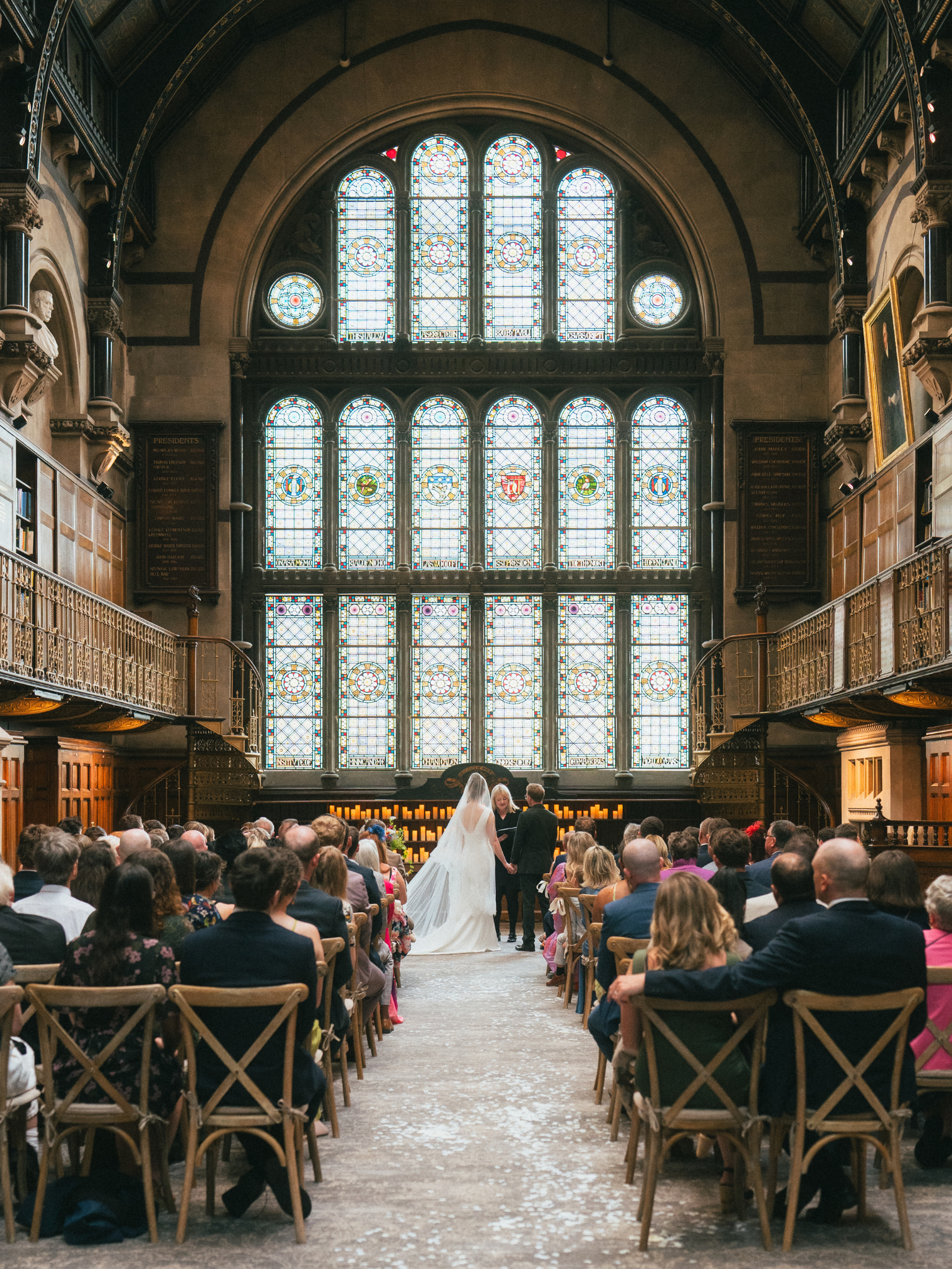 A wedding ceremony inside a grand church with large stained glass windows, a bride and groom holding hands at the altar, surrounded by seated guests.