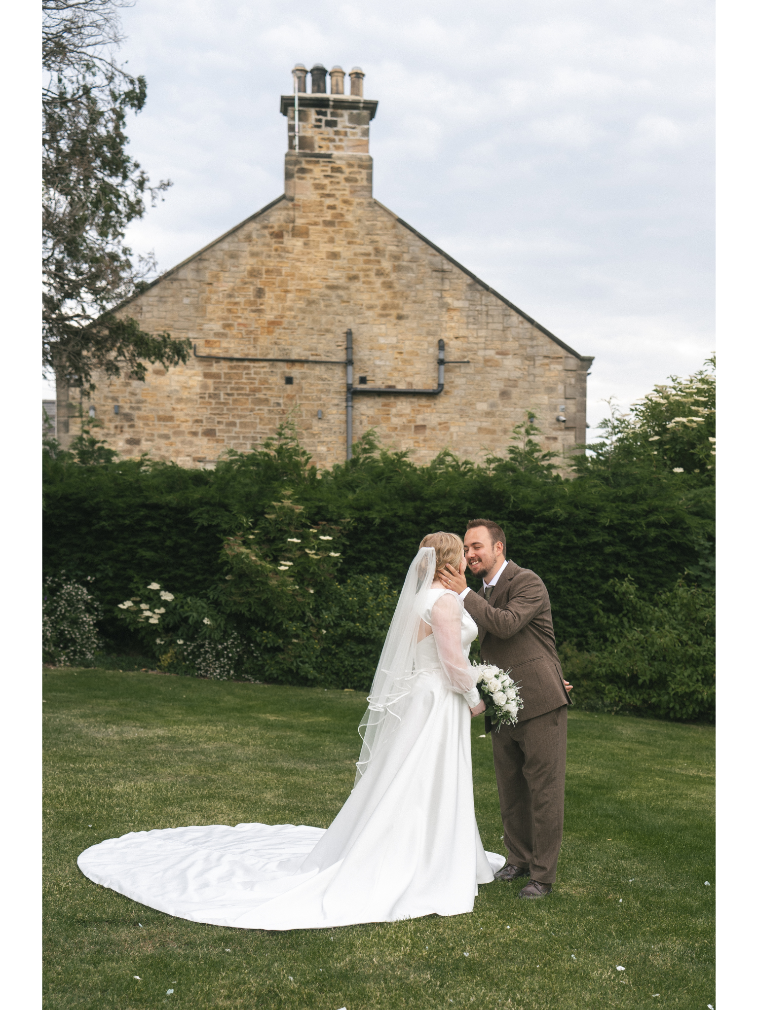 A bride and groom are standing outdoors on a grassy lawn, holding hands and smiling at each other. The bride is wearing a white wedding dress with a long train and veil, carrying a bouquet of white flowers. The groom is dressed in a brown suit and ti