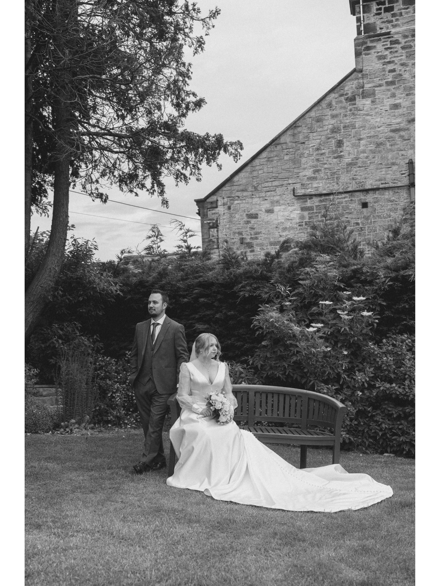 A black and white photo of a bride sitting on a park bench with a bouquet, while a groom stands beside her in what appears to be a garden setting with trees, shrubs, and a brick building in the background.