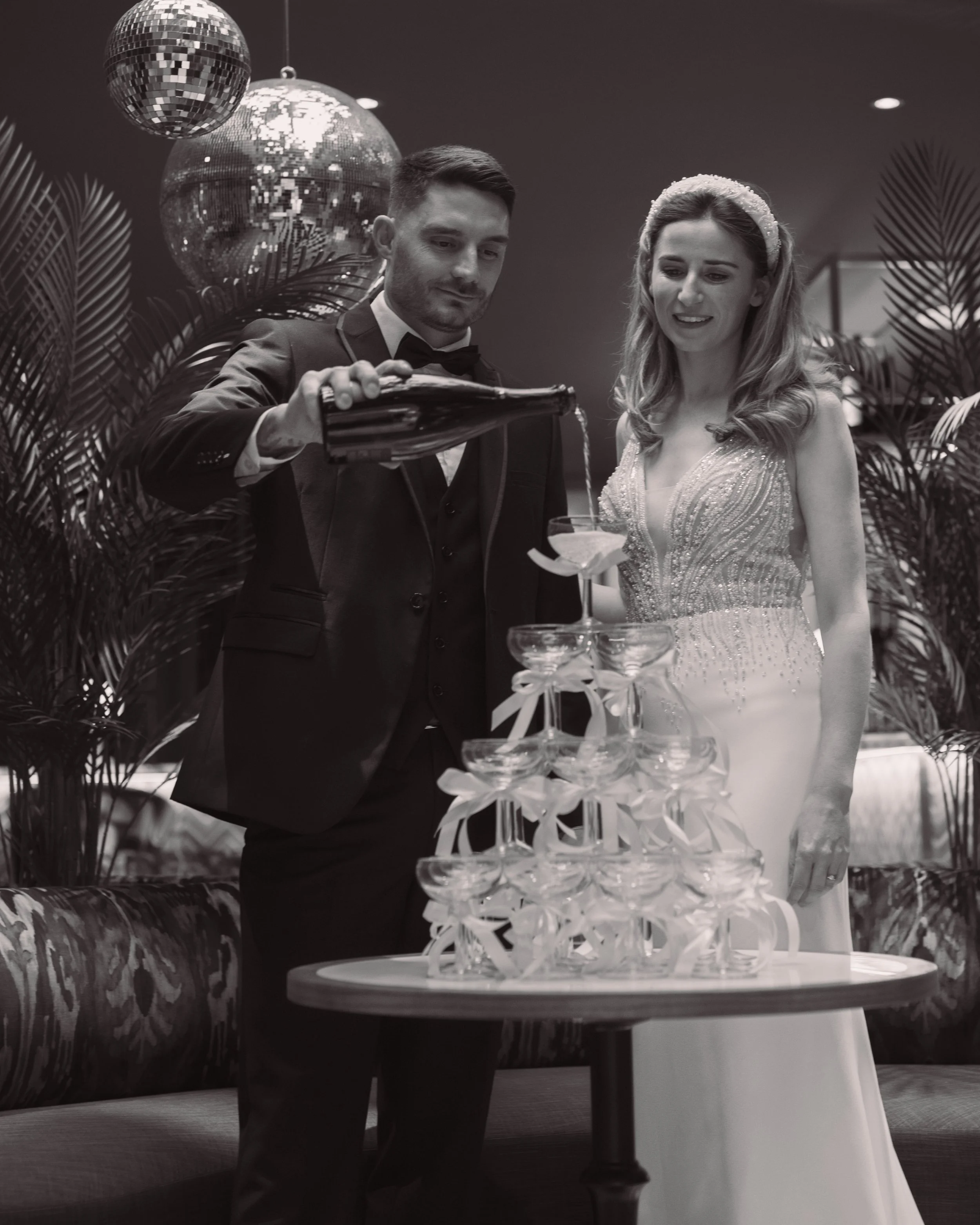 Black and white photo of a couple in formal attire pouring champagne into a tower of glasses at an event, with disco balls hanging overhead.