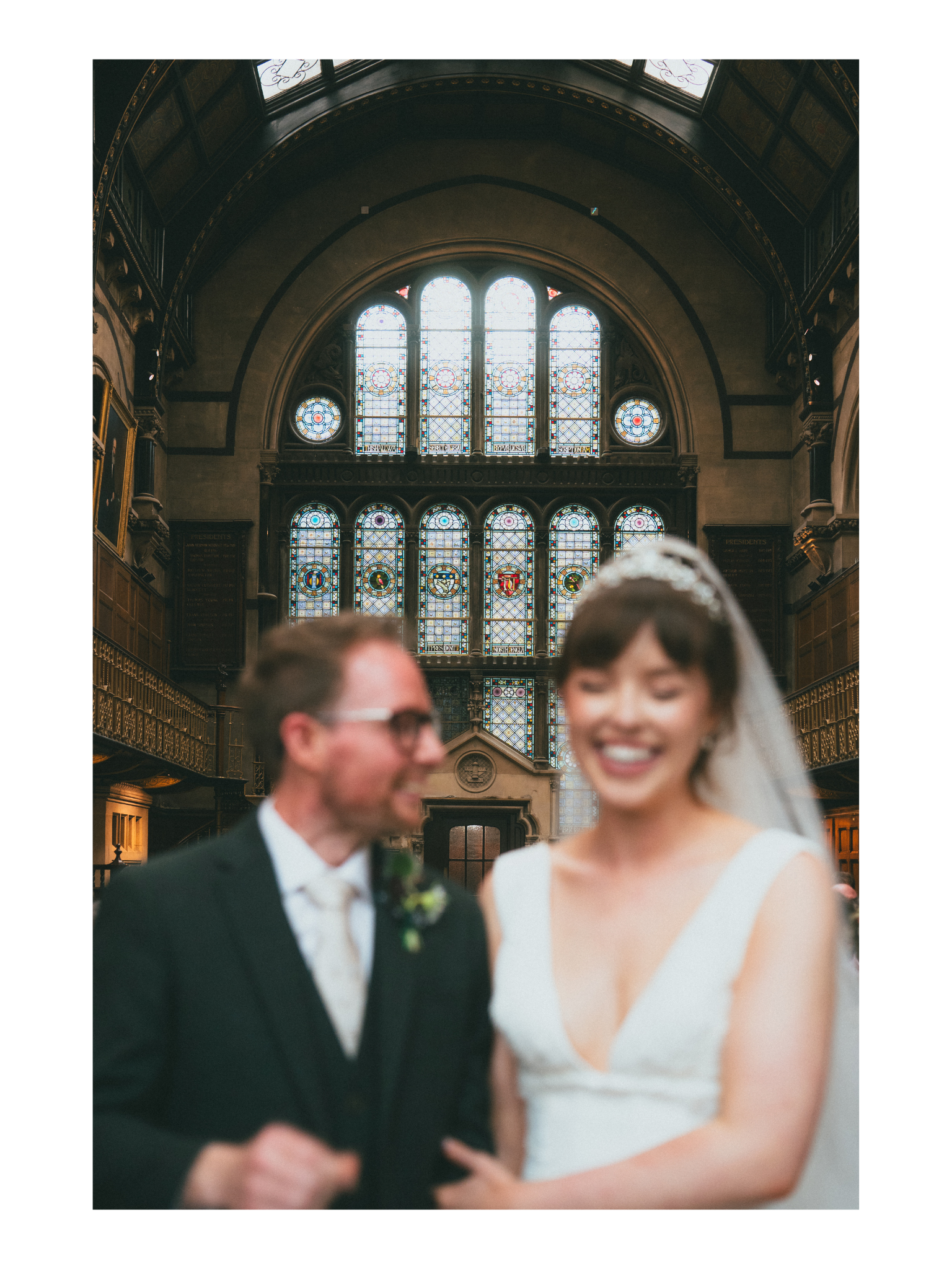 A wedding ceremony inside a historic building with stained glass windows, showing a smiling bride in a white dress and a groom in a dark suit, slightly blurred in the foreground.