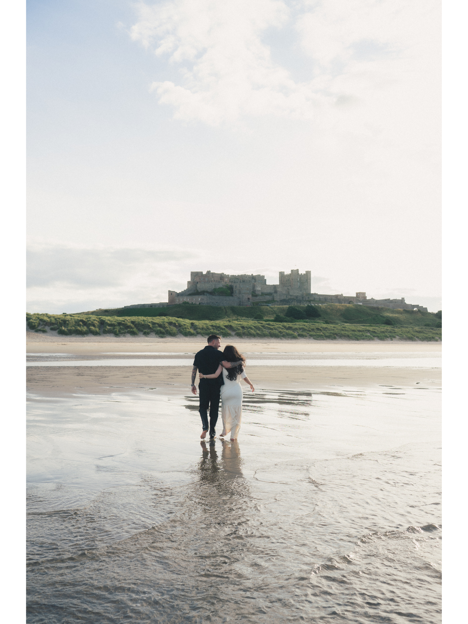 A couple walking on the beach with an ancient castle on a hill in the background during daytime.