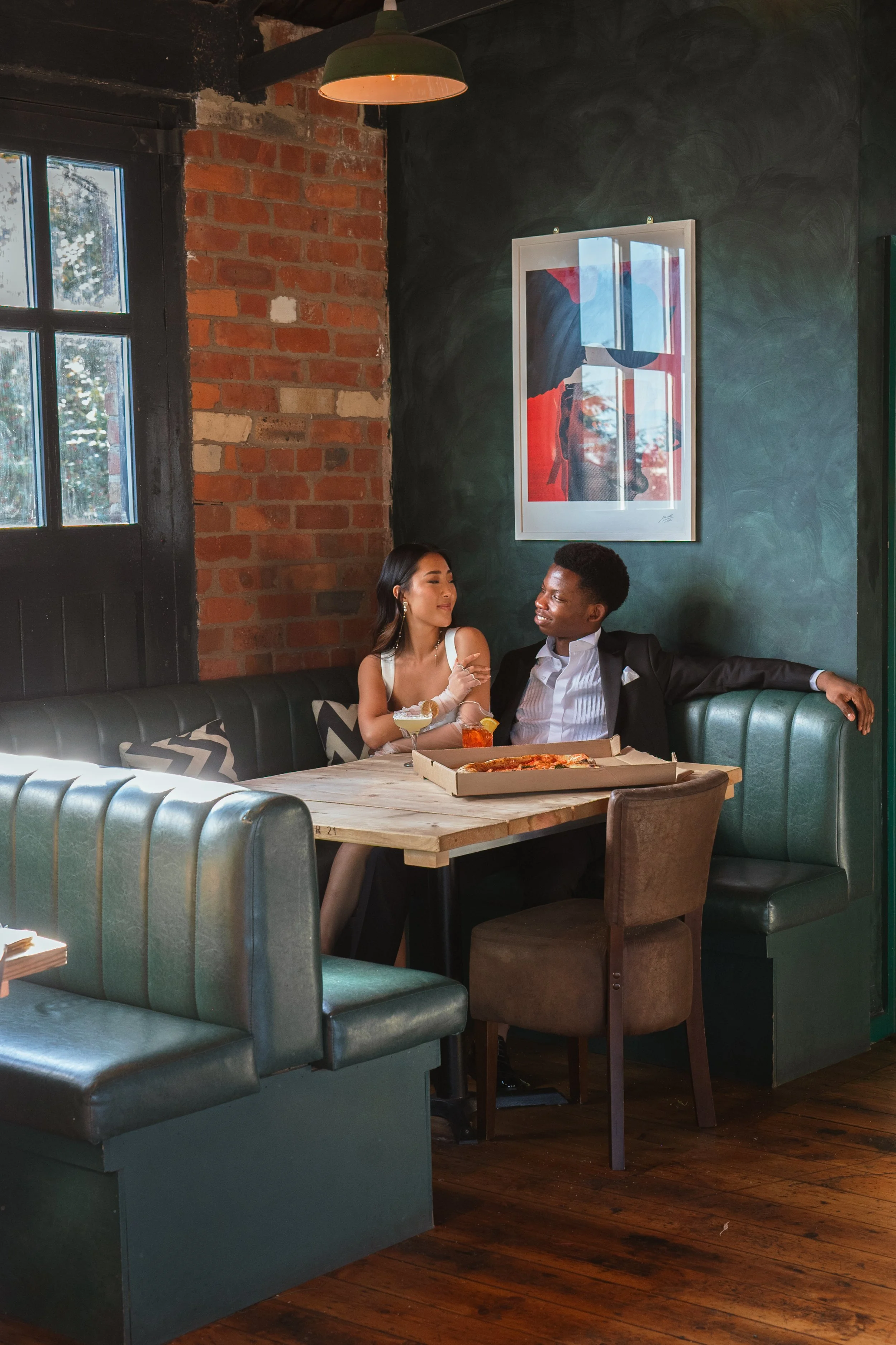 Couple sitting in a cozy restaurant booth, sharing a meal with pizza on a wooden table, brick and green walls, and abstract art on the wall.