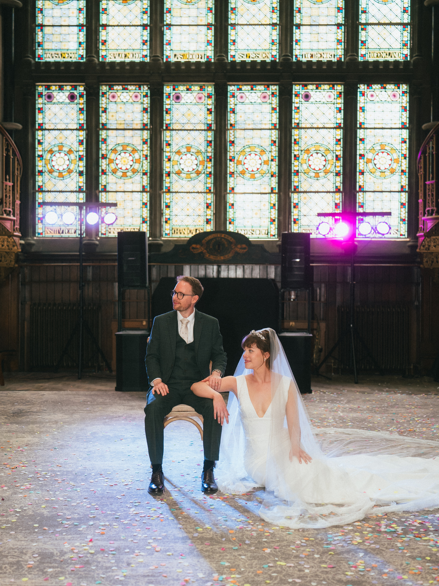 A bride and groom are sitting together in a large hall with stained glass windows at the back. The bride is wearing a white wedding dress with a veil, and the groom is dressed in a dark suit with glasses. The floor is scattered with colorful confetti