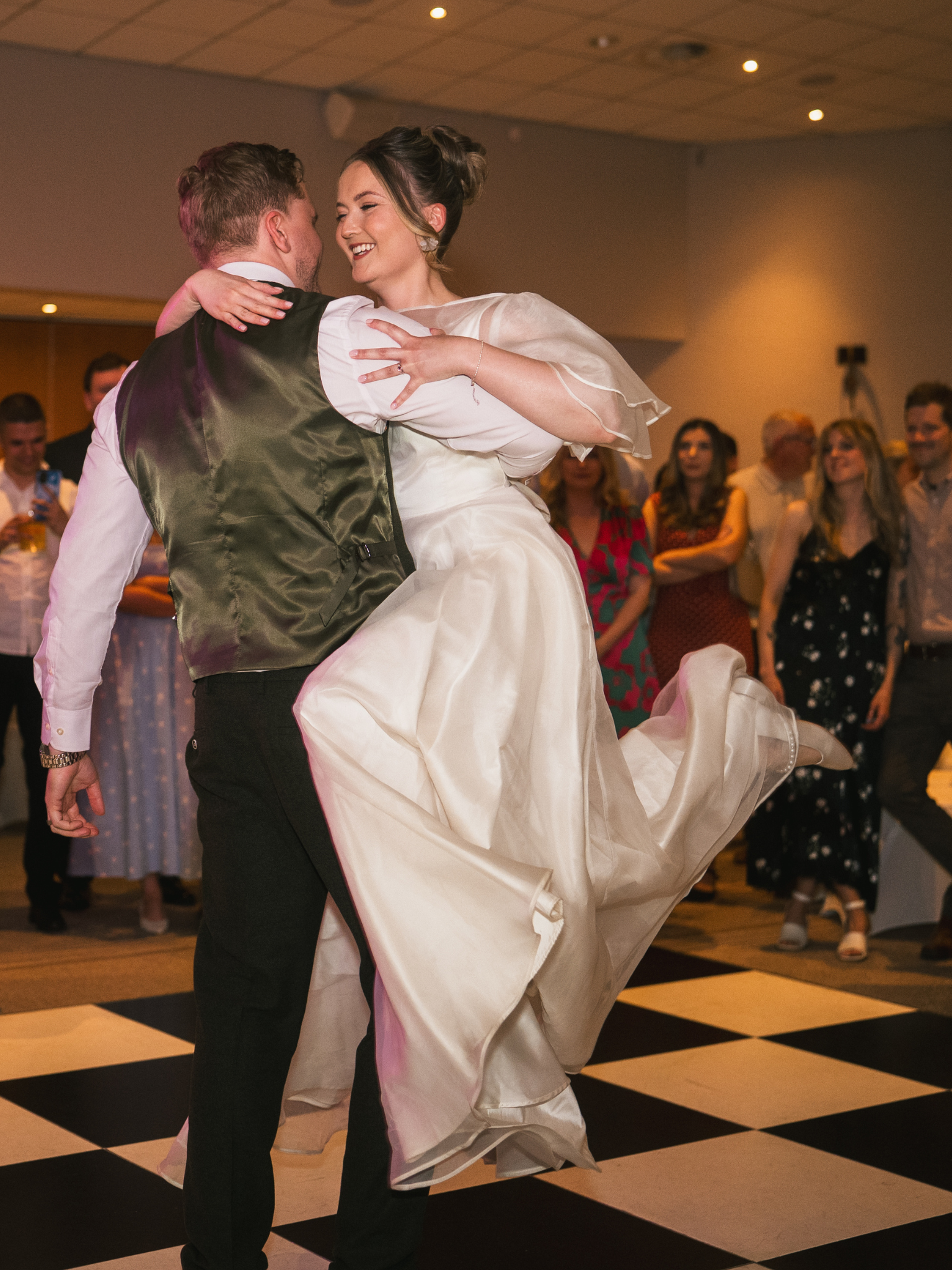 A couple dancing at a wedding reception, with the bride wearing a white gown and the groom in a vest, dancing closely together while guests watch and smile in the background.