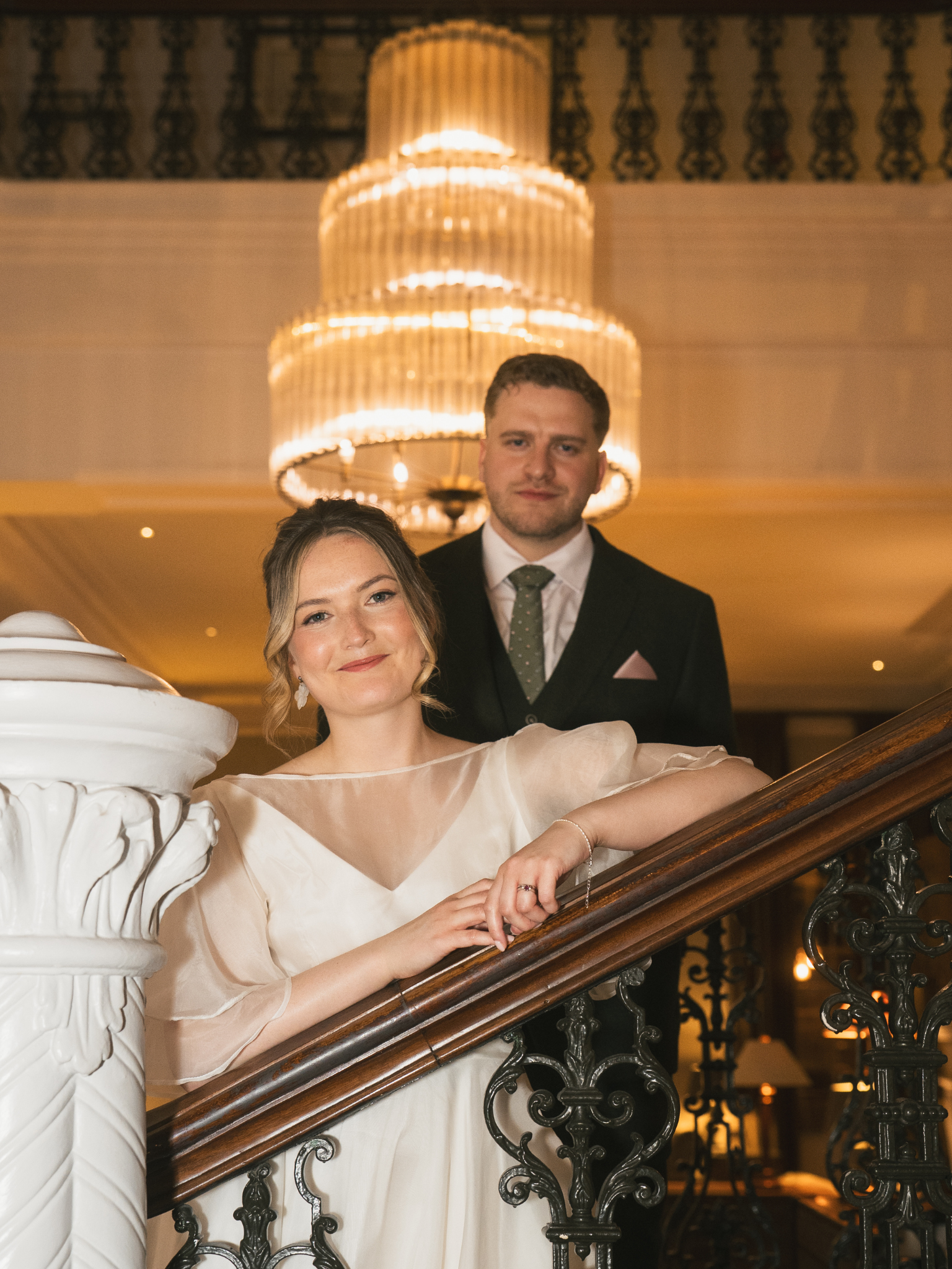 A couple posing for a photo on a staircase in an elegant setting. The woman, dressed in a light-colored dress with sheer sleeves, is smiling and resting her arm on the staircase railing. The man in a dark suit with a patterned tie stands behind her, 