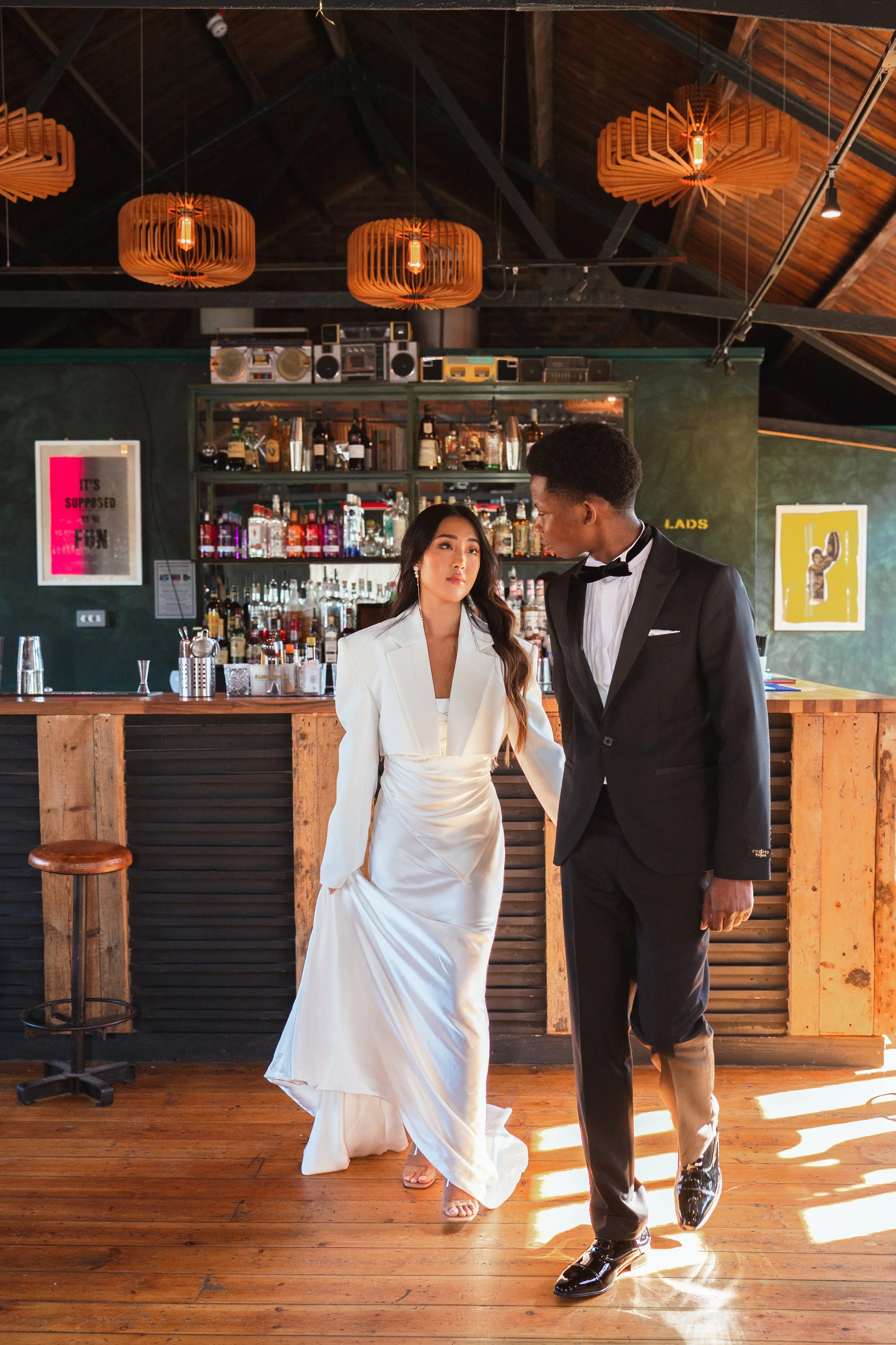 Couple in formal attire walking inside a bar with wooden flooring and vintage decor.