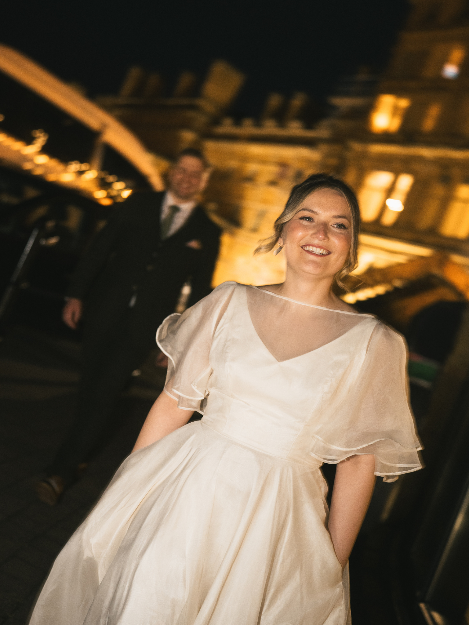 A smiling young woman in a cream-colored dress at a dimly lit event, with a man in a suit blurred in the background.
