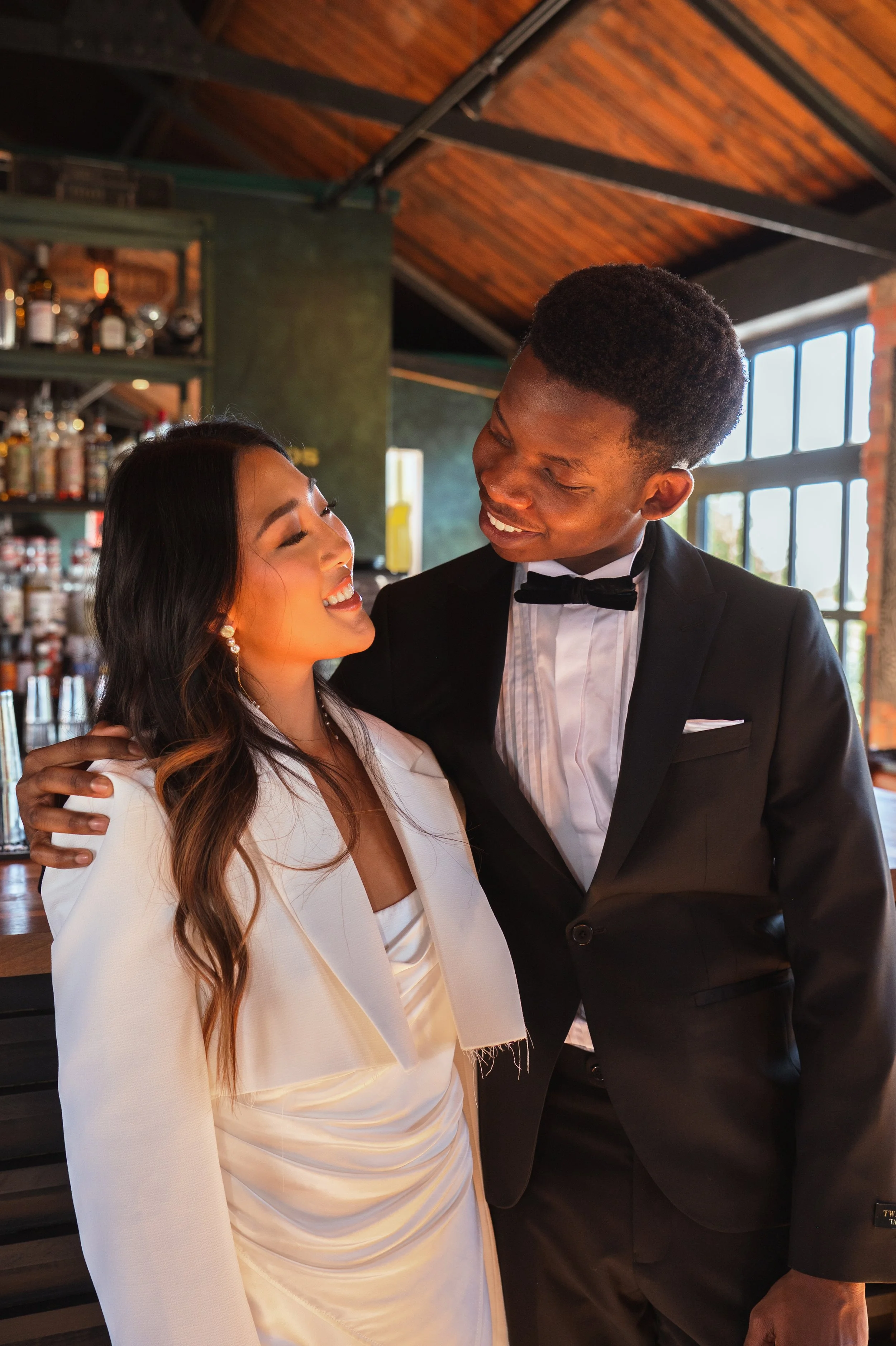 A couple in formal attire standing in a warmly lit room with a bar in the background. The woman is wearing a white dress and jacket, while the man is in a black tuxedo with a bow tie.