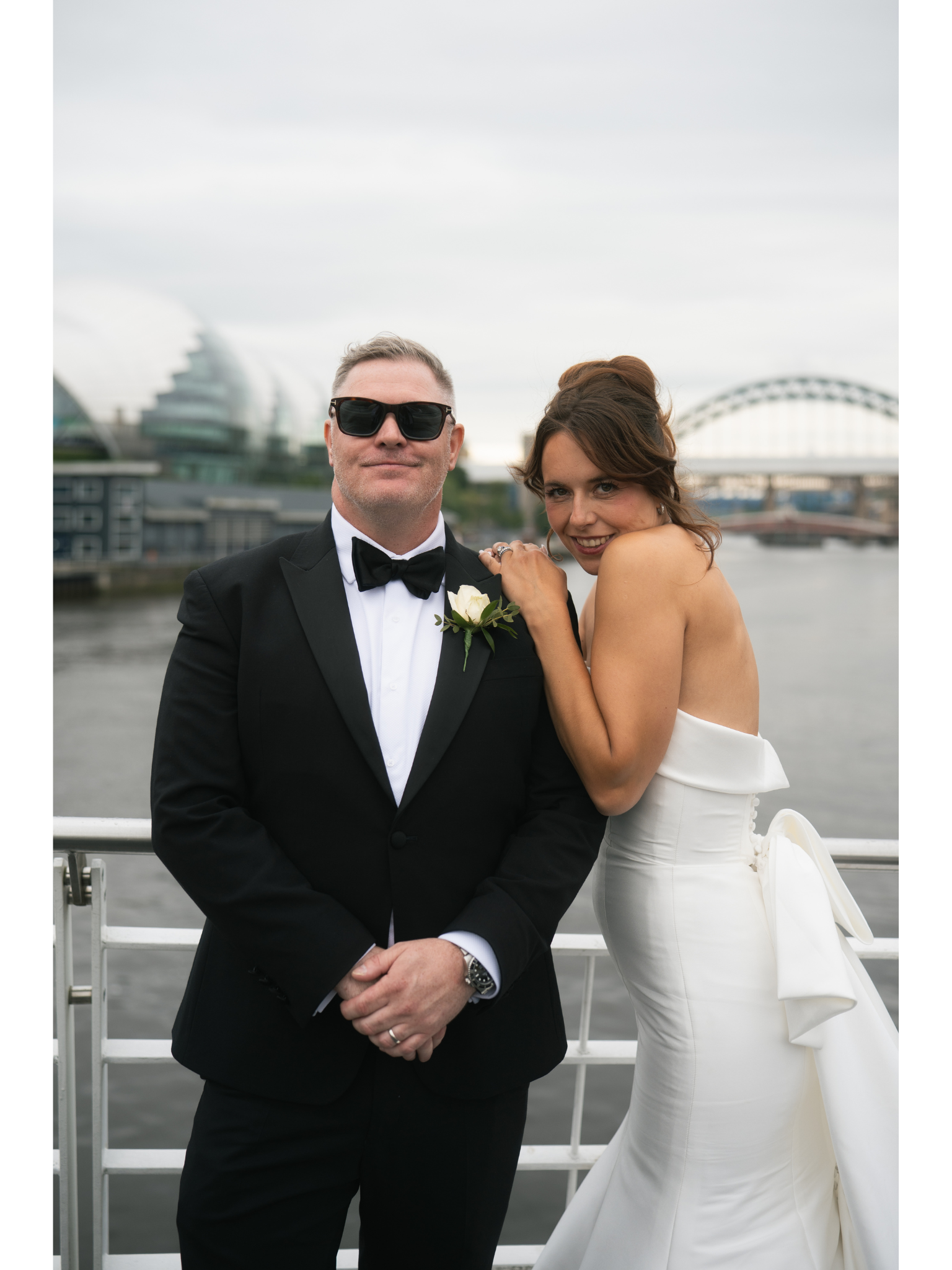 A couple dressed in wedding attire on a boat with a city skyline, including a modern glass building and a bridge, in the background.