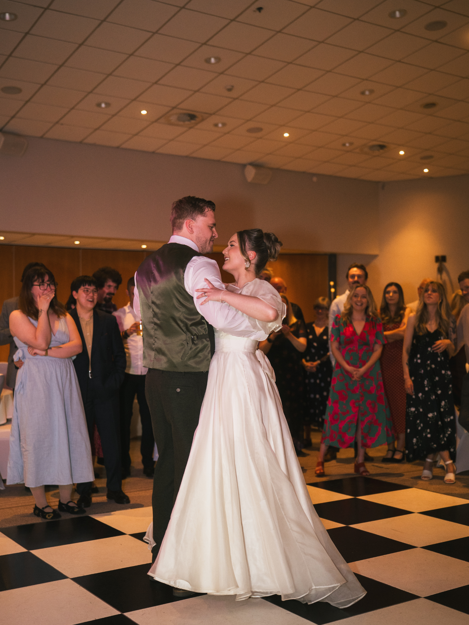 A bride and groom are dancing together at their wedding reception, surrounded by guests who are watching and smiling.